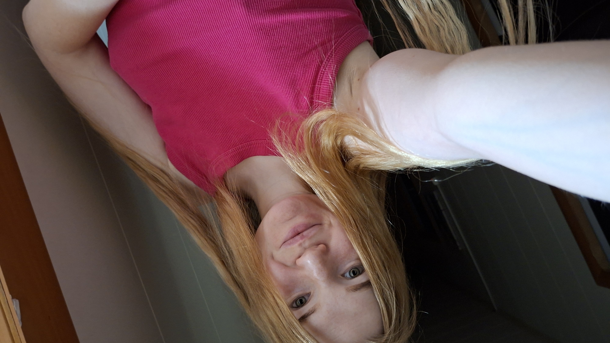 A close-up, selfie-style portrait features a young individual, likely female, with fair skin and long, light blonde hair. She is wearing a bright pink, ribbed short-sleeved top. The person is looking directly at the camera with a gentle, calm smile. The image is oriented with the subject's head towards the lower right and her body extending towards the upper left, suggesting she might be lying down or holding the camera at an unusual angle. Part of her arm is visible in the foreground. The background indicates an indoor setting, with muted tones of grey and brown walls, possibly showing vertical paneling and a dark, indistinct area. No text is visible in the image.