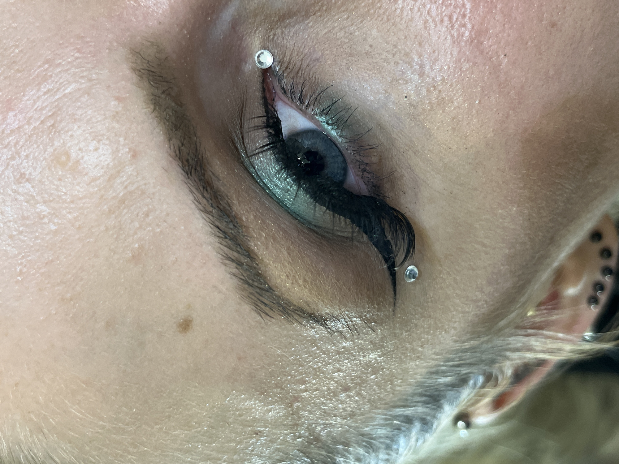 A close-up photograph shows a person's eye, featuring makeup, false eyelashes, and two small, silver-colored studs positioned above and below the eye near the tear duct. The person has fair skin with a few visible blemishes. The eye is partially open, revealing a dark iris and distinct eyelashes. There are other piercings visible in the ear and scattered on the face. The image is not connected to the city of Plovdiv, Bulgaria.
