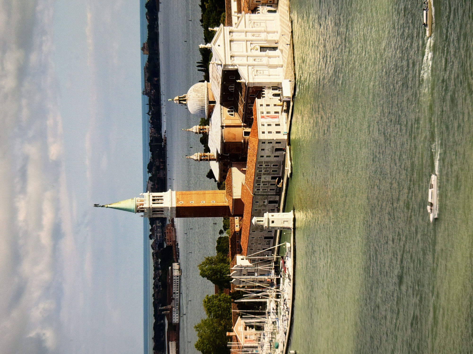 This is an aerial, sideways view of a waterfront city, likely Venice, Italy, given the architectural style and canal system. The scene is dominated by a large body of water, appearing green and slightly choppy, suggesting a breezy day. Several white boats, including a sleek, modern yacht, are visible on the water.
On the land, there are numerous historic buildings with red-tiled roofs and white facades. Prominent structures include a large white church with a dome and several towers, one of which is tall and brick with a green, spire-like top. The architecture suggests a European, possibly Renaissance, influence. Trees are interspersed among the buildings, adding a touch of greenery to the urban landscape. The sky is a pale blue with soft, white clouds.
The image appears to be taken during the daytime, likely late morning or early afternoon, as the lighting is bright but diffused. There is no discernible activity of people, beyond the boats on the water. No text is clearly visible in the image. The overall impression is one of historical grandeur and serene maritime life. The provided location context of Kyiv, Ukraine, is inconsistent with the visual evidence, which strongly points to Venice.