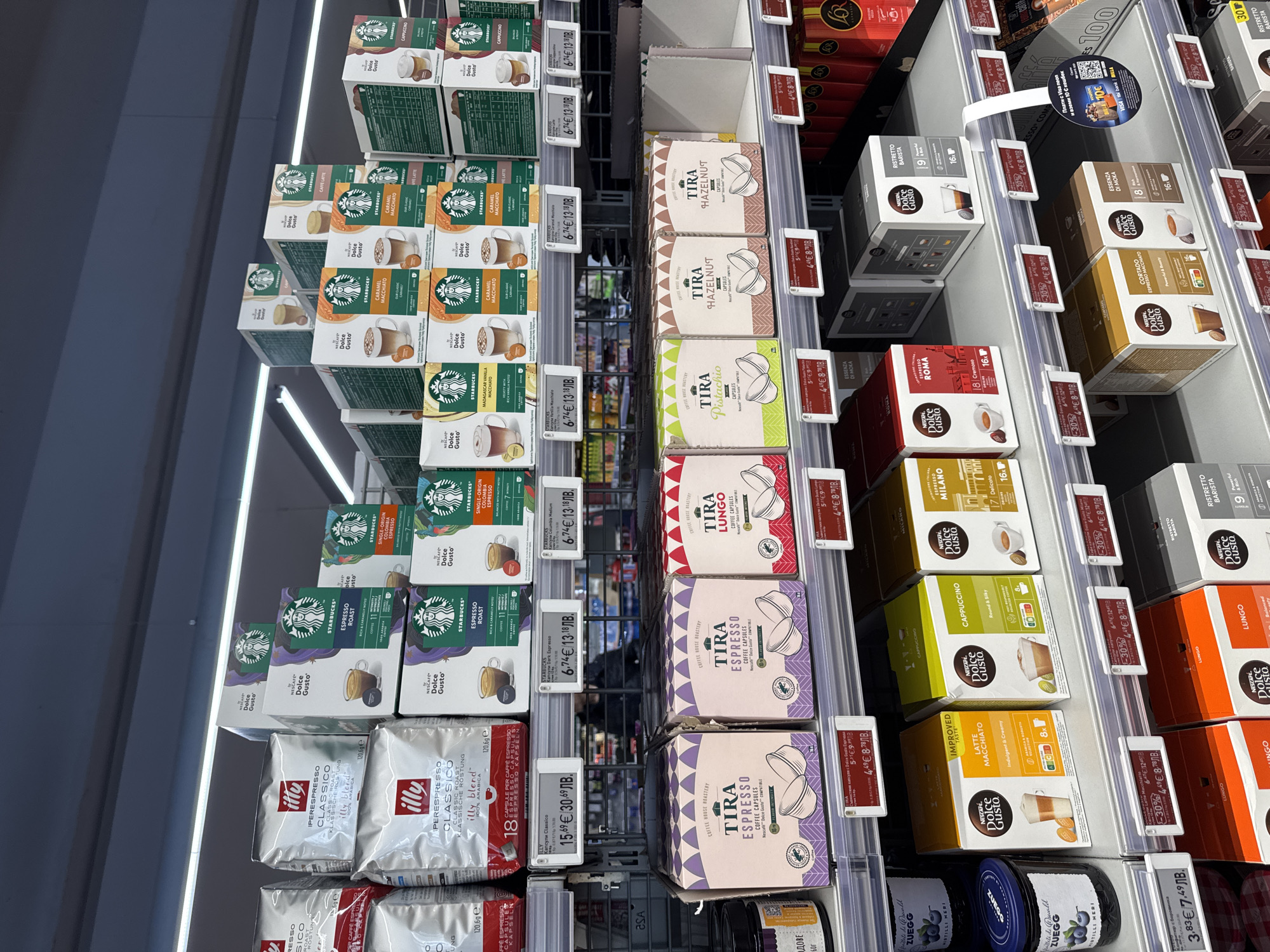 The image shows a shelf in a store in Sofia, Bulgaria. The shelf is stocked with various coffee capsules, including brands like Starbucks, Illy, and Nescafé Dolce Gusto. The capsules are arranged in rows with price tags. Other items like jams are also visible on the shelves.