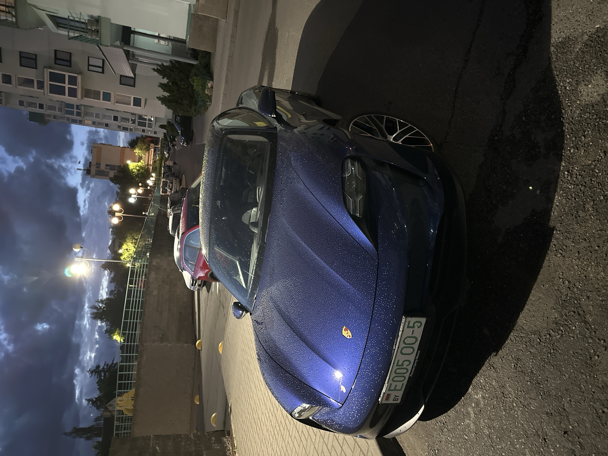 The image depicts a wet, blue Porsche Taycan parked on a brick-paved area next to a street. The car's license plate reads "E005 00-5 BY," indicating it is registered in Belarus, and the text "Porsche Центр Минск" (Porsche Center Minsk) is also visible on the plate. The car is parked near a low wall or barrier with yellow markings, and a few other vehicles are parked in the distance. The sky is overcast with dark clouds. The scene appears to be in a residential or commercial area of Minsk, Belarus, based on the architecture of the buildings in the background and the license plate information. The lighting suggests it is dusk or evening. No people are visible.