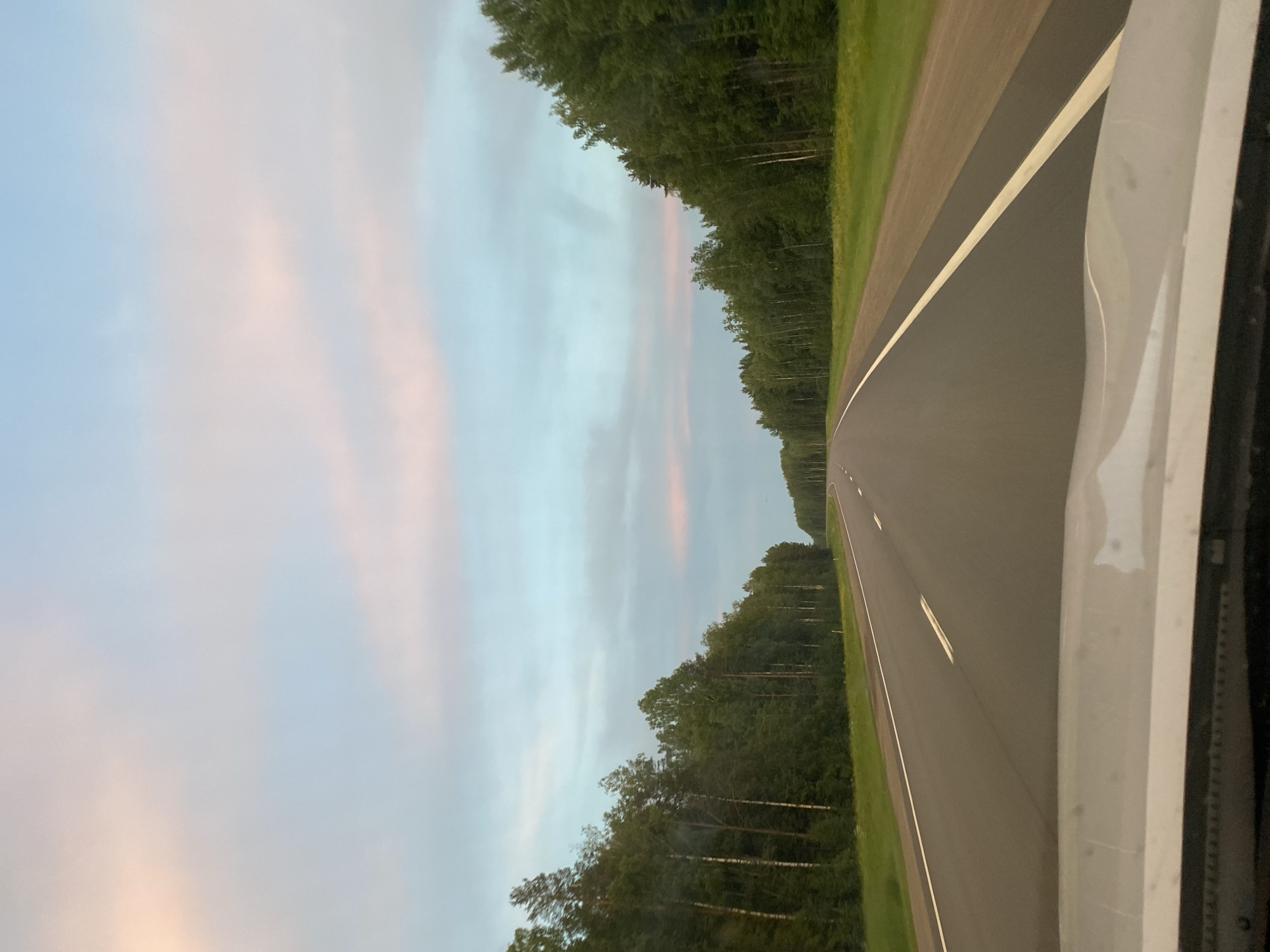 The image depicts a road trip through a natural landscape, likely on the outskirts of a city. The perspective is from inside a vehicle, looking out through the windshield. The road is paved and extends into the distance, with white lane markings visible. On either side of the road, dense green forests line the path. The sky above is a soft blend of blue and pink, suggesting either dawn or dusk. There are no people or buildings immediately visible in the frame, emphasizing the natural and open setting. The location is identified as the city of Первомайский, Russia.