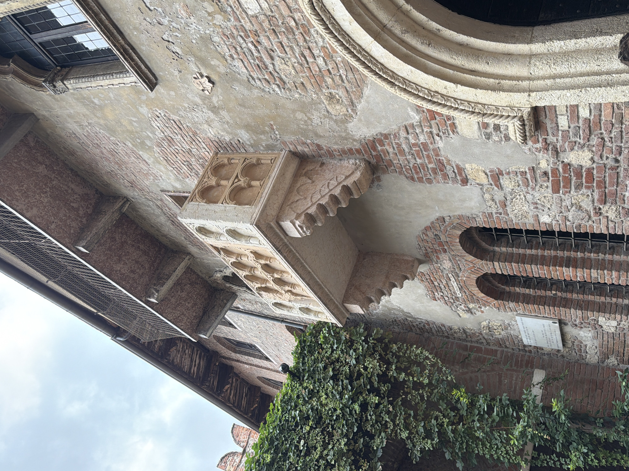 The image shows the exterior of a historic building in Verona, Italy, famously known as Juliet's House. A prominent stone balcony, adorned with Gothic-style tracery, juts out from the brick facade. Green ivy cascades down the left side of the building, partially obscuring arched windows with iron grilles. The weathered brickwork shows signs of age, with some areas of plaster missing. A decorative arched doorway is visible on the right, featuring a rope-like stone molding. The sky above is overcast. There are no people clearly visible in the frame, though a small portion of a person's head and shoulders is visible in the lower right corner. A plaque is mounted on the wall near the ivy.
