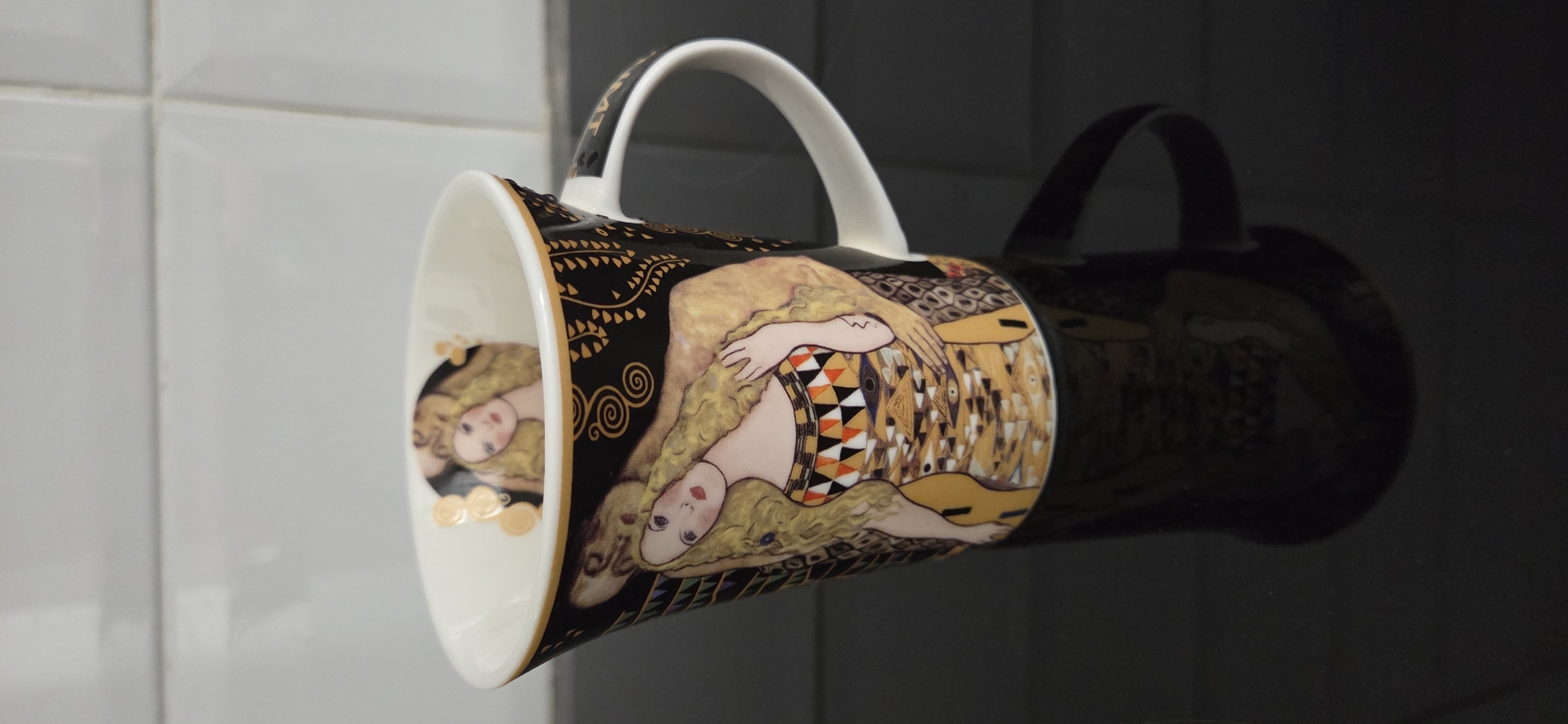 A tapered ceramic mug featuring an Art Nouveau-inspired design, predominantly black with gold accents, is positioned centrally on a highly reflective dark surface. The mug's exterior displays a stylized female figure with long, flowing golden hair and a geometrically patterned dress in hues of gold, black, white, and orange, set against a black background with intricate gold swirls and linear patterns. The interior of the mug is white, with a smaller, similar female figure depicted on its inner base. The mug has a white handle on its right side and a thin gold band along its rim. A clear, inverted reflection of the mug, including its design and handle, is visible on the dark surface below. In the background, a light-colored tiled wall with visible grout lines forms a grid pattern. No specific location, such as the city of Lipetsk, Russia, can be determined from the visual information provided in the image.