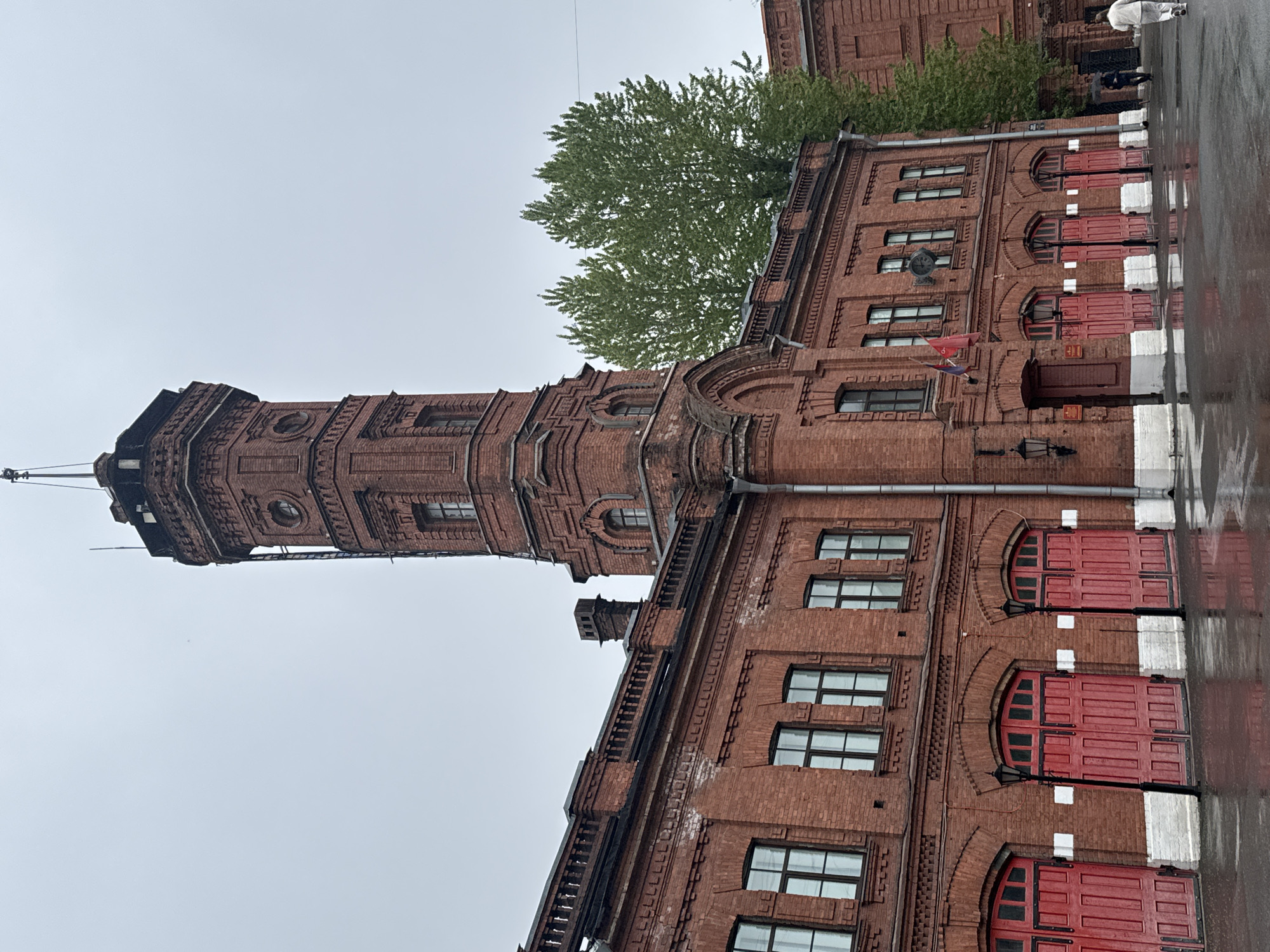 A low-angle, vertically-oriented shot depicts a historic brick building with a prominent tower on a cloudy day. The building is likely a fire station, given the multiple red doors that appear to be large vehicle access points. The tower is richly ornamented with brickwork, and its top is capped with a spire. Windows are visible throughout the building, and there are two flags hanging from the side of the building. A tree partially obscures the upper portion of the building. The ground appears to be wet, reflecting parts of the building. The overall scene suggests a location in the city of Saint Petersburg, Russia, based on the architectural style.