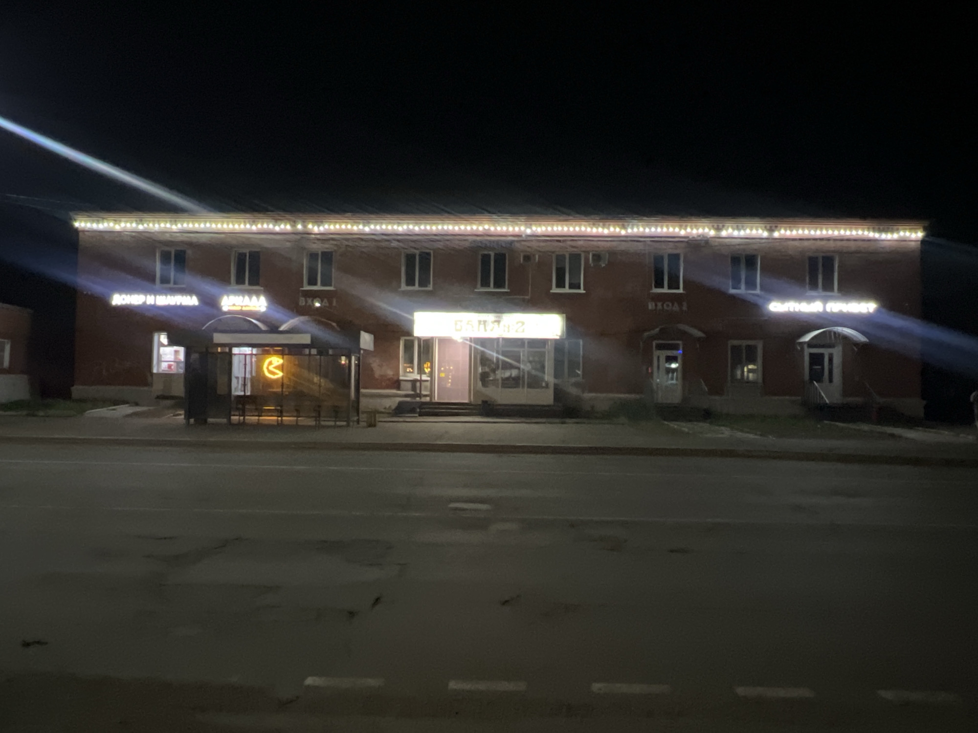 The image shows a two-story brick building at night in what is likely Chaikovsky, Russia. The building is decorated with lights along the roofline. There are several signs in Russian above the entrances, including "ДОНЕР И ШАУРМА", "АРКАДА", "БАНЯ-2", and "СЫТЫЙ ПРИВЕТ". One entrance has a neon sign depicting a Pac-Man-like figure. In front of the building is a road with visible patches. There are no people visible in the image. Light streaks emanate from the signs and streetlights.