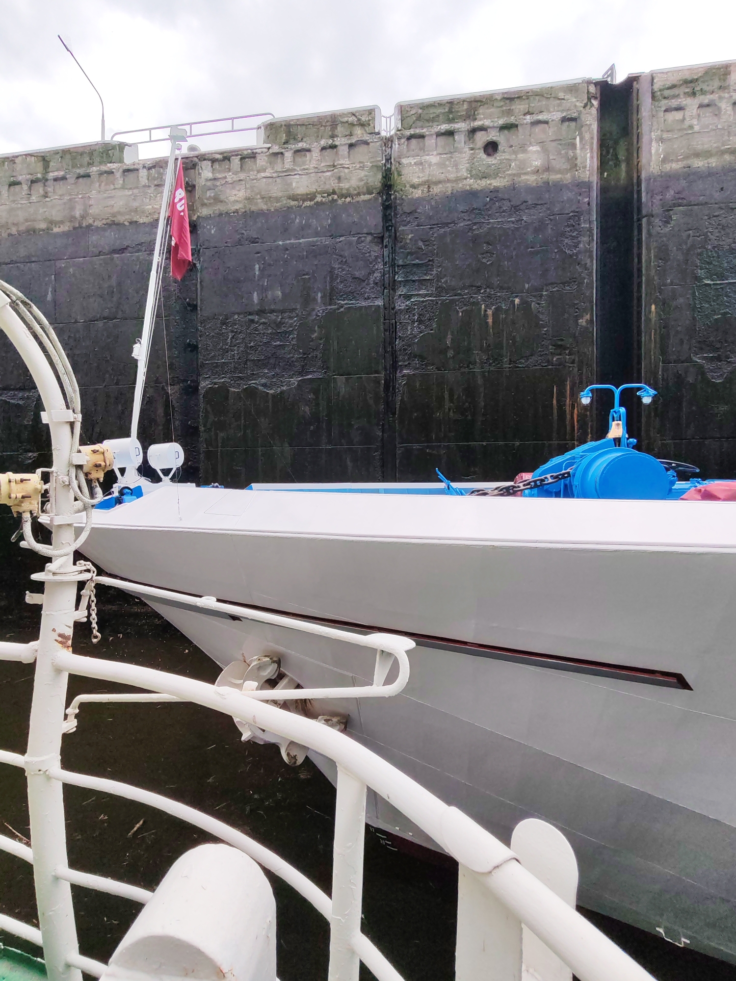 In the image, a white boat is positioned in a lock on a waterway. The lock gates, made of dark, rough stone, are visible in the background, with a gap indicating the lock is in operation or transitioning. A red flag, with some white text, flutters from a pole on the boat. The boat itself has a white hull with a blue trim. A portion of another boat, with white railings, is in the foreground, suggesting the perspective of someone on a vessel within the lock or adjacent to it.
The scene appears to be outdoors, under an overcast sky, suggesting a cloudy or possibly rainy day. The water in the lock is dark and appears murky, with some debris floating on the surface. The stonework of the lock walls shows signs of wear and possibly algae or moss growth, indicating it's an established structure.
The overall impression is of a functional, industrial waterway environment. There are no people clearly visible in the frame. The activity taking place is likely the process of locking the boat through the waterway, either entering or exiting the lock. The red flag might signify the nationality or registration of the boat. The white text on the flag is partially obscured but appears to be in Cyrillic. The location context provided is Bol'shie Mkhi, Russia, which aligns with the possibility of a waterway lock system.