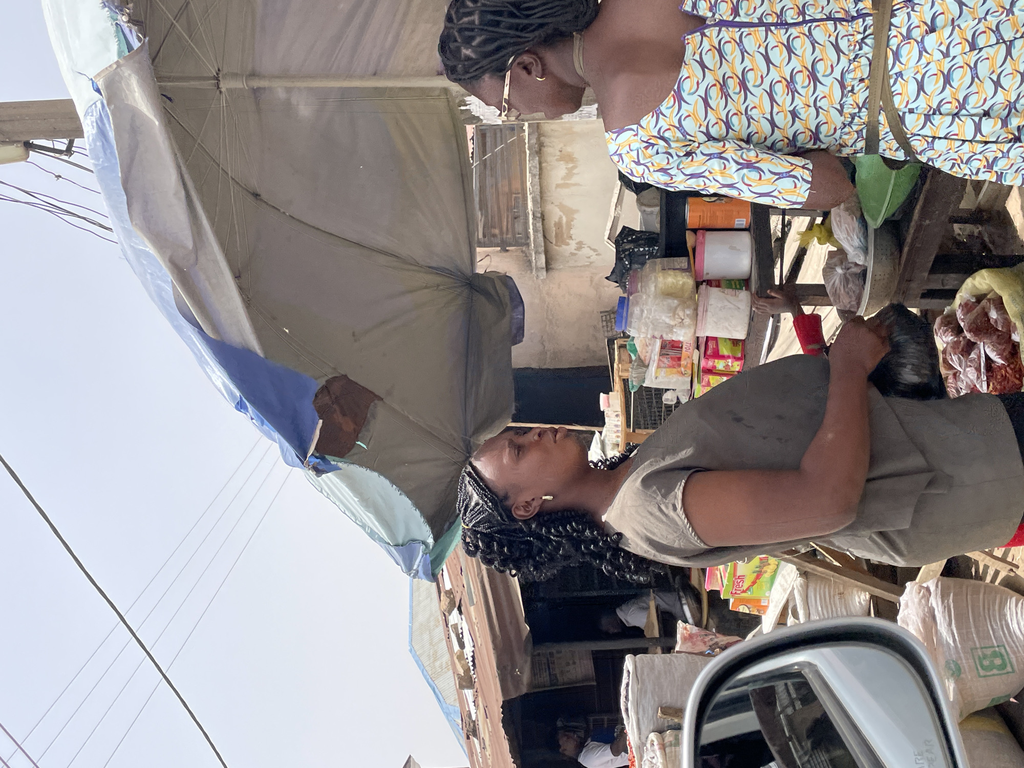 The image depicts an outdoor market scene in Jos, Nigeria. A woman with braided hair, wearing a neutral-colored top, is engaged in a transaction at a market stall. The stall, covered by a makeshift awning of grey and blue tarpaulin, displays various goods. Another woman, adorned in a patterned blue and yellow dress, stands at the stall as well. The market environment includes crowded stalls, with items like food and bags visible. The image is taken from the perspective of someone inside a car, with the car's side mirror visible in the foreground.