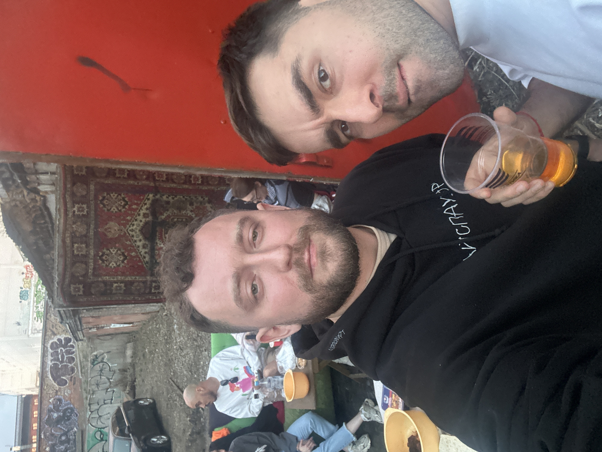 Two men are holding a plastic cup and taking a selfie. The photo includes other people in the background. The location is Krasnoyarsk, Russia, as suggested by the markings on the cup. A worn carpet is displayed on the wall. Graffiti is visible on a nearby building.