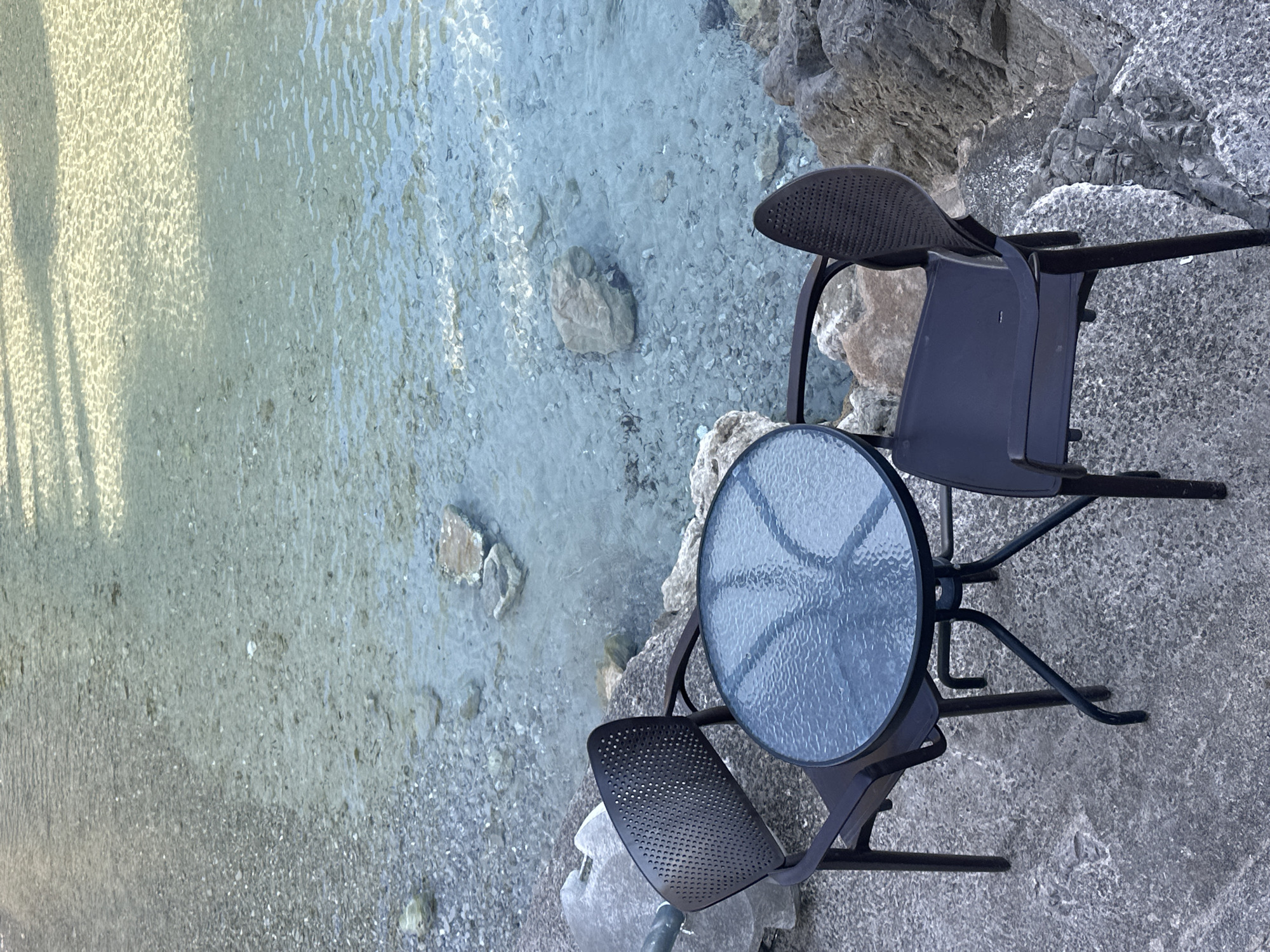 The image depicts a serene outdoor setting by a body of water in Seoca, Montenegro. In the immediate foreground, a rough, grey ground surface, consistent with concrete or paved stone, anchors the scene. Positioned on this surface are two dark-colored, possibly black or dark brown, outdoor chairs with perforated backs and a small, round table with a dark metal frame and a textured, translucent glass top. One chair is situated to the left of the table, angled towards it, while the second chair is on the right, also angled towards the table. No individuals are present at or near the table.
Beyond the seating arrangement, the terrain transitions into a rocky shoreline composed of varying sizes of grey and brown stones. This leads into a body of exceptionally clear, shallow water. The water's surface is lightly rippled, reflecting sunlight in various patterns, and its transparency allows for an unobstructed view of the underwater environment. The bottom is covered with numerous small, light-colored pebbles and several larger, irregular rocks. Distinct shadows are cast across the upper section of the water, suggesting overhead structures or foliage not visible within the frame. The overall impression is one of tranquility and natural beauty.