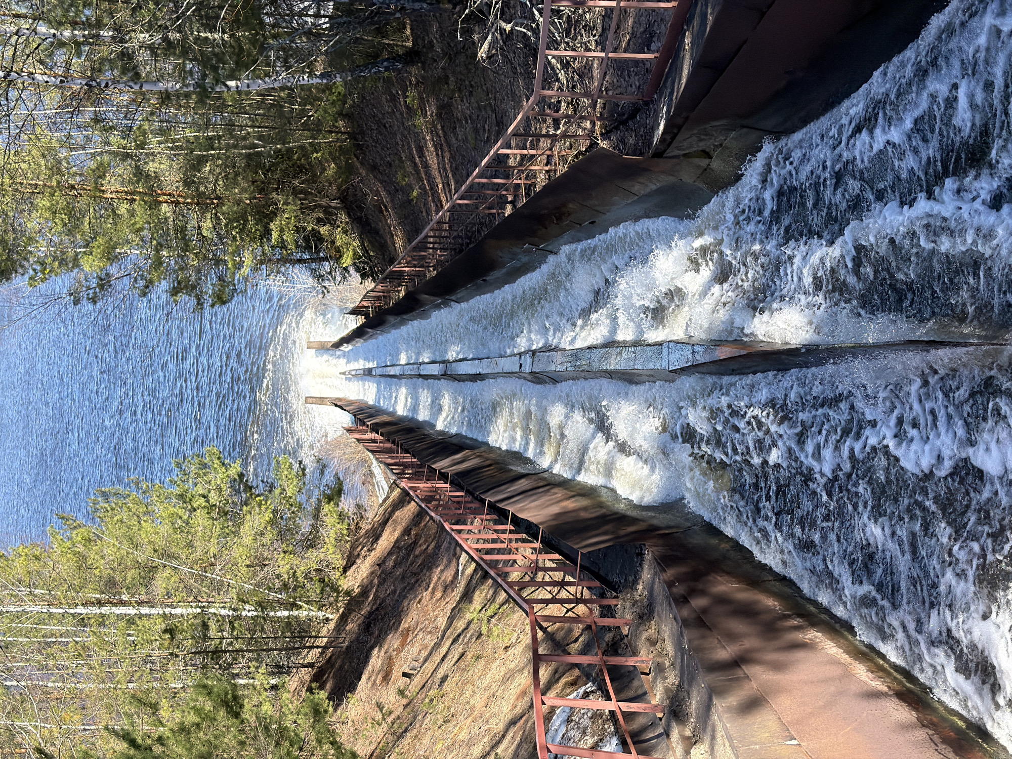 The image depicts a water chute in Chernushka, Russia. The chute is concrete with metal railings on either side. Water cascades down the chute towards a larger body of water. Trees are visible on both sides of the chute. The bank adjacent to the chute appears grassy with some dry vegetation. The lighting suggests a sunny day.