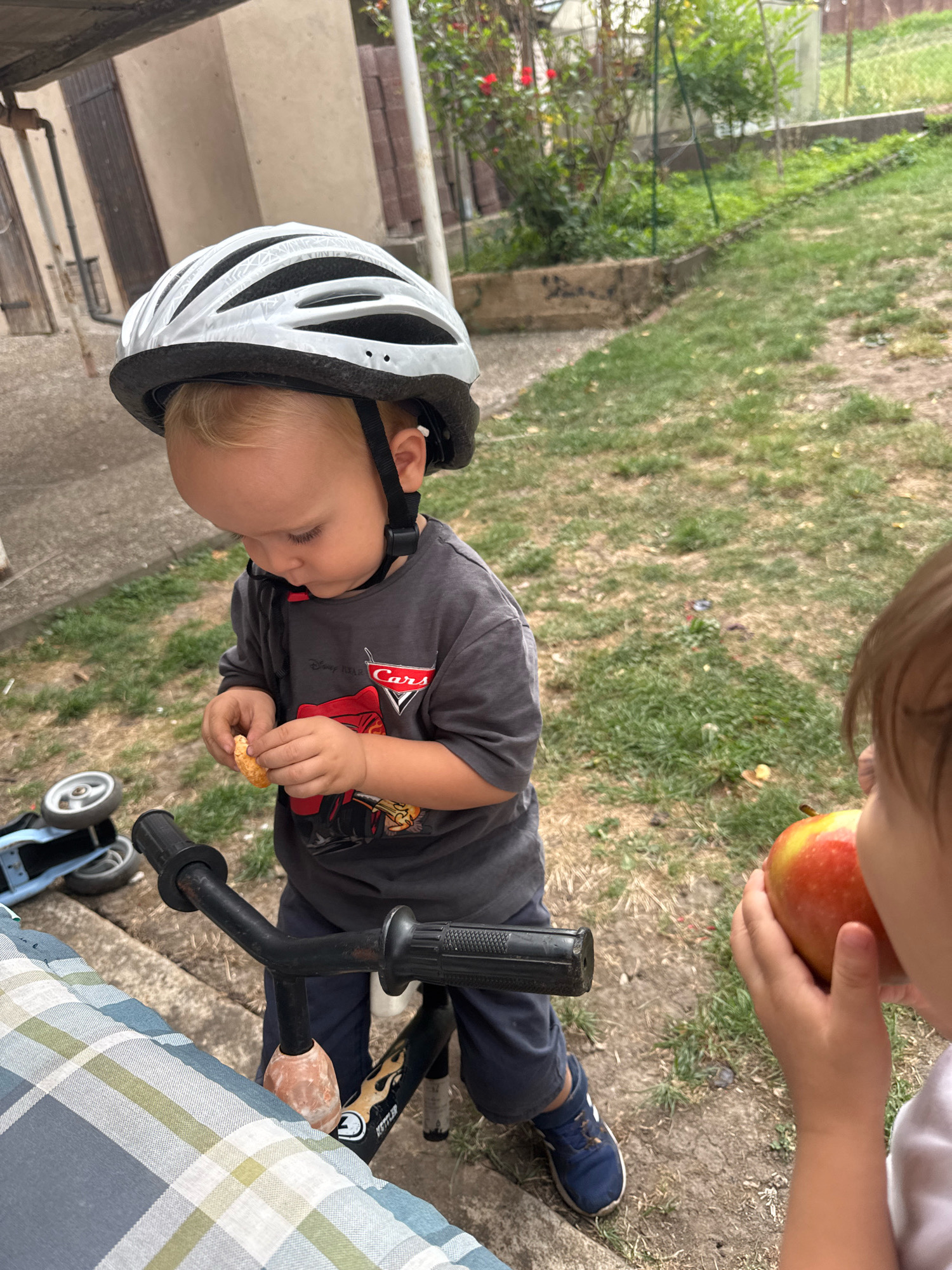 The image depicts a young child wearing a white and black bicycle helmet, a grey t-shirt with a Disney Pixar Cars graphic, and dark pants. The child is holding a small piece of food in their hands and appears to be looking down at it. To the right of the child, another child is visible, holding a red and yellow apple. The scene appears to be an outdoor setting, possibly a backyard, with grass, dirt, and some vegetation in the background. A bicycle with training wheels is partially visible in the foreground. The location is identified as Überherrn, Germany.