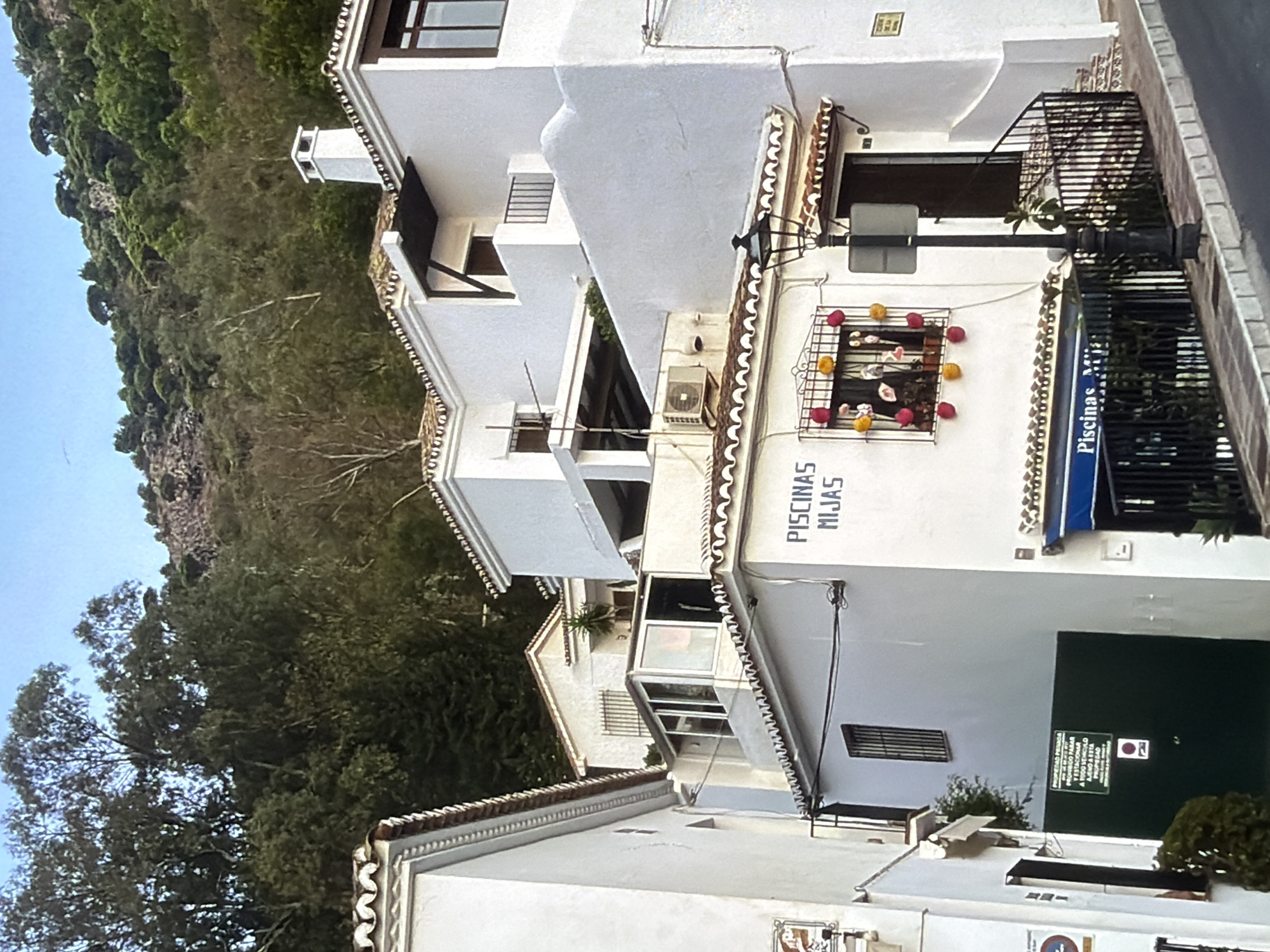 This is a photograph of a street scene in a European village. The buildings are whitewashed with terracotta roof tiles, characteristic of Mediterranean architecture. A steep, tree-covered hillside rises behind the buildings. The sky is clear and blue. The image appears to be taken during daylight hours, likely late afternoon or early evening, given the shadows.
The foreground shows part of a street with a dark asphalt surface and a brick sidewalk. A dark metal street lamp with a decorative flourish stands on the sidewalk. In the background, white buildings with dark-trimmed windows and balconies are stacked closely together. One building has a sign with blue lettering that reads "PISCINAS MIJAS" which translates to "MIJAS POOLS". Another building features a decorative window grill with colorful ornaments, possibly red and yellow pom-poms or flowers. Above the street, several stories of the white buildings are visible, including chimneys and window air conditioning units. A small, yellow sign is affixed to the wall of one building.
The scene appears to be quiet, with no people visible. The overall impression is one of a charming, possibly tourist-oriented, village. The vegetation on the hillside is dense and green, suggesting a mild climate.