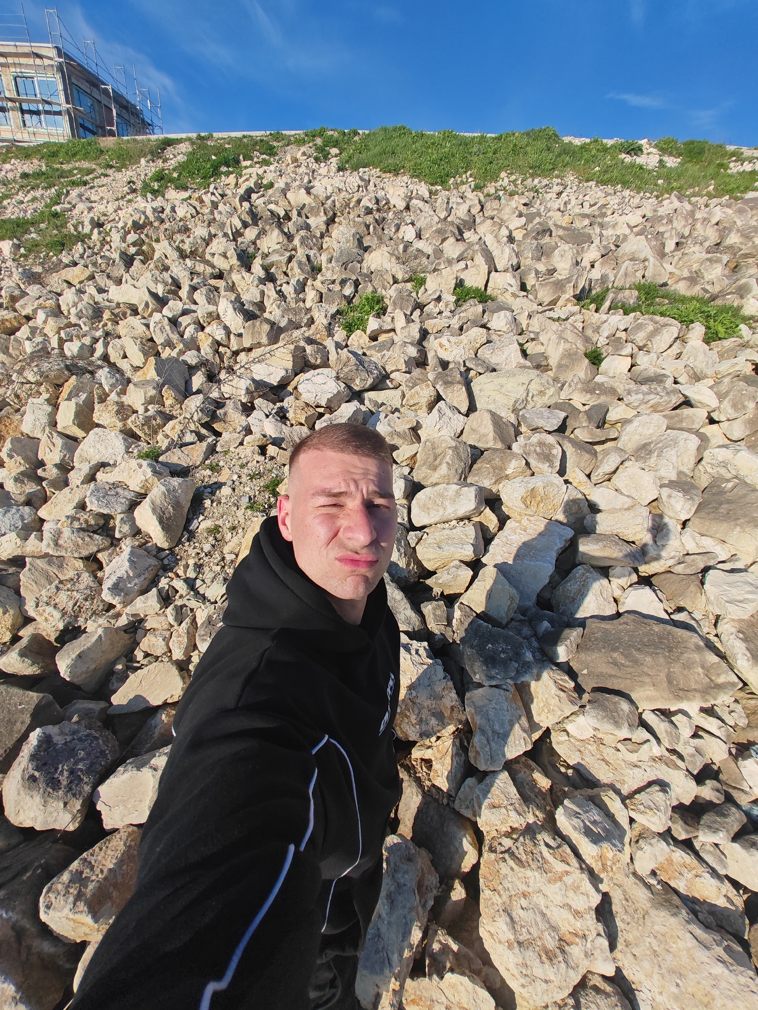 A man is captured in a selfie on a steep, rocky hillside under a clear blue sky. He is positioned in the lower-left to central part of the frame, looking directly at the camera with a slightly scrunched, pensive, or perhaps mildly annoyed expression, his mouth slightly pursed. He has short, light-colored hair and is wearing a black hooded sweatshirt (hoodie) with some white detailing or stripes visible on the sleeves and body. His black athletic pants also feature prominent white stripes running down the side of the legs.
The vast majority of the image is dominated by the hillside, which is heavily covered with numerous irregularly shaped, light-colored rocks, appearing to be limestone or a similar type of stone. These rocks vary in size, from small pebbles to larger boulders, and are interspersed with sparse patches of vibrant green grass and weeds, particularly along the upper sections of the slope. The incline is quite steep, suggesting a challenging terrain.
In the upper-left background, at the crest of the hill, a building under construction is visible. It features multiple windows and is surrounded by scaffolding. Beyond the hill, a broad expanse of bright, clear blue sky with only a few faint wisps of white clouds fills the upper right portion of the frame. The scene is brightly lit by direct sunlight, casting distinct shadows from the rocks and the man onto the ground, indicating either morning or late afternoon. The overall impression is one of rugged outdoor scenery, possibly near a coastal or newly developed area. No discernible text is visible in the image.