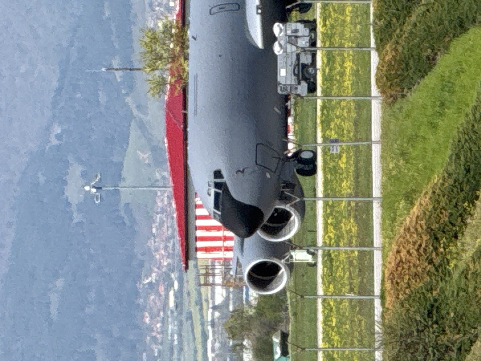 An aerial view of an airplane, most likely a cargo plane or military transport, parked on a grassy area or runway. The aircraft is painted in a dark grey or black color, with its nose pointing towards the viewer. Two large engines are visible beneath the fuselage near the front. A structure with red and white stripes is located near the tail of the aircraft. Behind the plane, a cityscape is visible, with buildings and possibly a mountain range in the background. In the foreground, a light green and brown grassy area is partitioned by what appear to be metal poles or fences. The location appears to be in the vicinity of Sofia, Bulgaria.