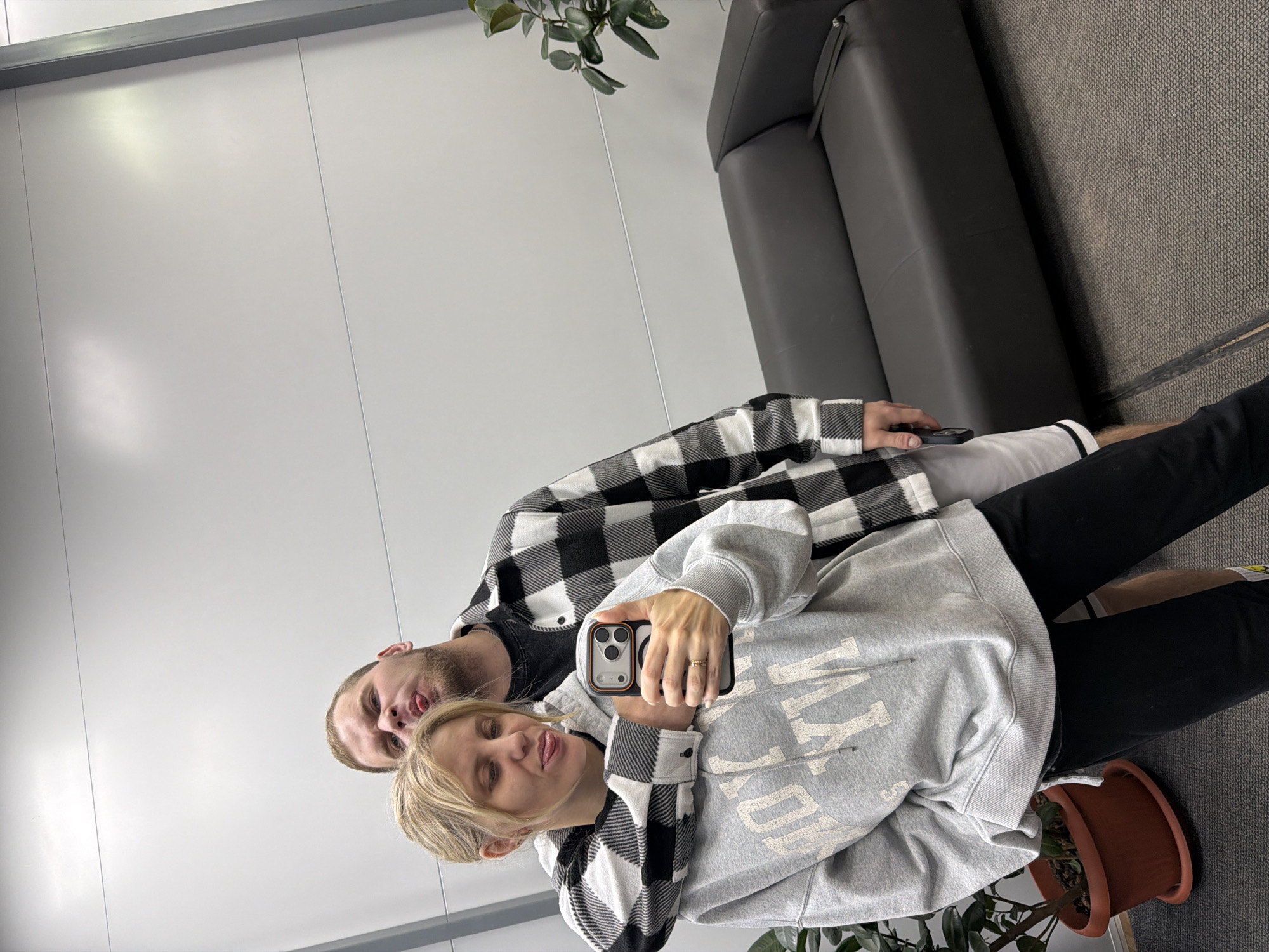 A man and a woman are captured in a selfie taken indoors. The image appears to be a casual moment, with both subjects making playful expressions, sticking out their tongues. The woman, with blonde hair, is in the foreground holding a smartphone to take the picture. She is wearing a grey hooded sweatshirt with white text and a black and white checkered jacket or shirt over it. A ring is visible on her left ring finger. The man, with short brown hair and a beard, stands behind her, also wearing a black and white checkered shirt over a dark t-shirt. He also has on light-colored shorts.
They are situated in what looks like a modern indoor space. The background features plain white walls and a dark grey sofa to the right. The floor is covered with a dark grey carpet. A potted plant is visible near the bottom right of the frame. The lighting is bright and even, consistent with an indoor setting.
Visible text on the woman's sweatshirt clearly shows "YORK" as part of a larger word or phrase, likely "NEW YORK". The overall atmosphere is lighthearted and informal.