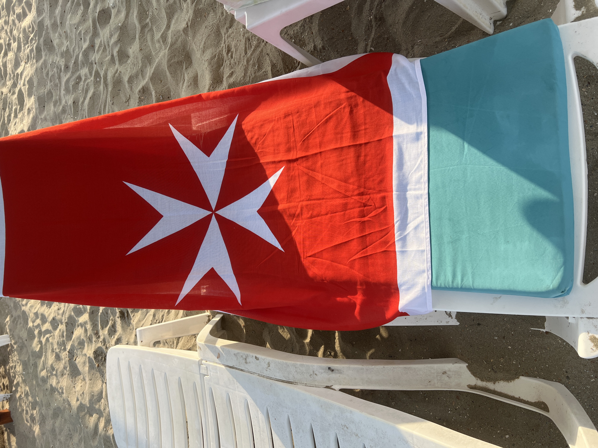 The image shows a close-up of a beach chair on a sandy beach. A red flag with a white Maltese cross is draped over the chair. The chair has a white plastic frame and a turquoise cushion. The sand is light brown and textured, with some shadows cast by the beach chairs. The scene appears to be in the city of Constanța, Romania, a popular seaside resort. There are no people or animals visible in the image. The lighting suggests it is either early morning or late afternoon, with the sun casting long shadows.