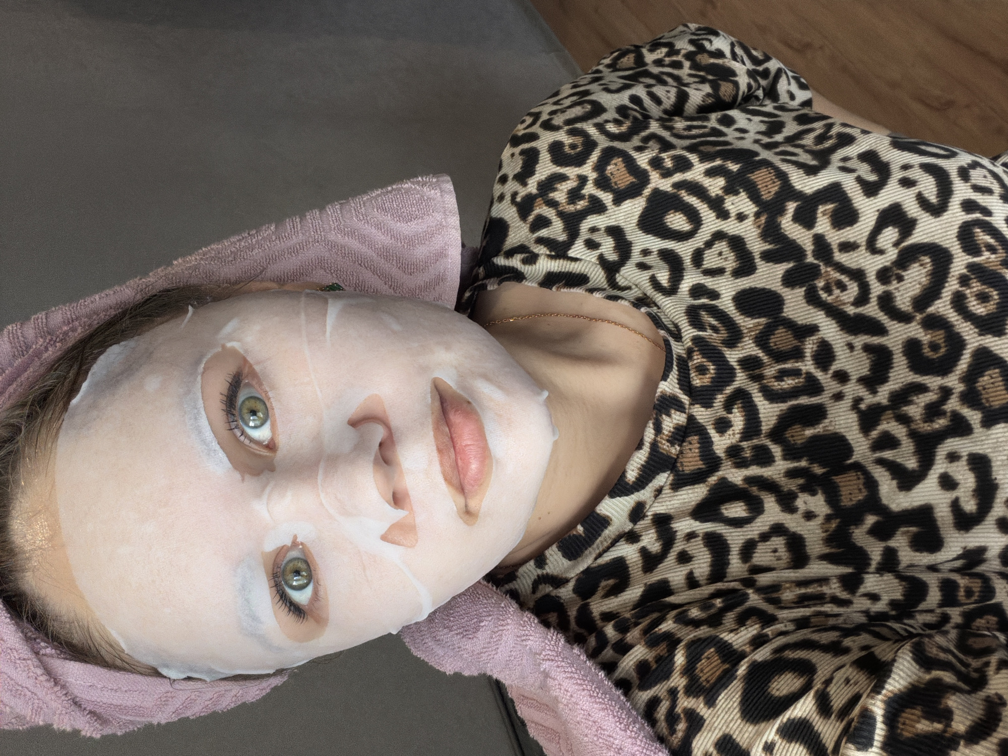 The image shows a close-up, overhead view of a person, likely a woman, lying down and wearing a sheet mask. Her head is tilted slightly to the left, and her gaze is directed upwards towards the camera.
**Subjects:** The primary subject is a person whose face is covered by a white sheet mask. Her eyes, which are a light green or hazel color, are visible through the mask's cutouts, as are her light pink lips. Her hair is covered by a light pink terry cloth towel wrapped around her head. She is wearing a top with a distinct leopard print pattern in shades of tan, brown, and black. A thin gold chain necklace is visible around her neck.
**Activity:** The person appears to be engaged in a skincare routine, specifically using a face mask, and is in a state of relaxation.
**Notable details:** The sheet mask is moist and fits closely to her facial contours. Her eyelashes are long and dark. The leopard print fabric of her clothing is textured, possibly ribbed. The background is mostly out of focus; a plain grey surface is visible behind her head, and a warm-toned wooden floor texture can be seen to the far right. The lighting is soft and even, suggesting an indoor setting. There is no visible text in the image. The time of day and specific location are not discernible.