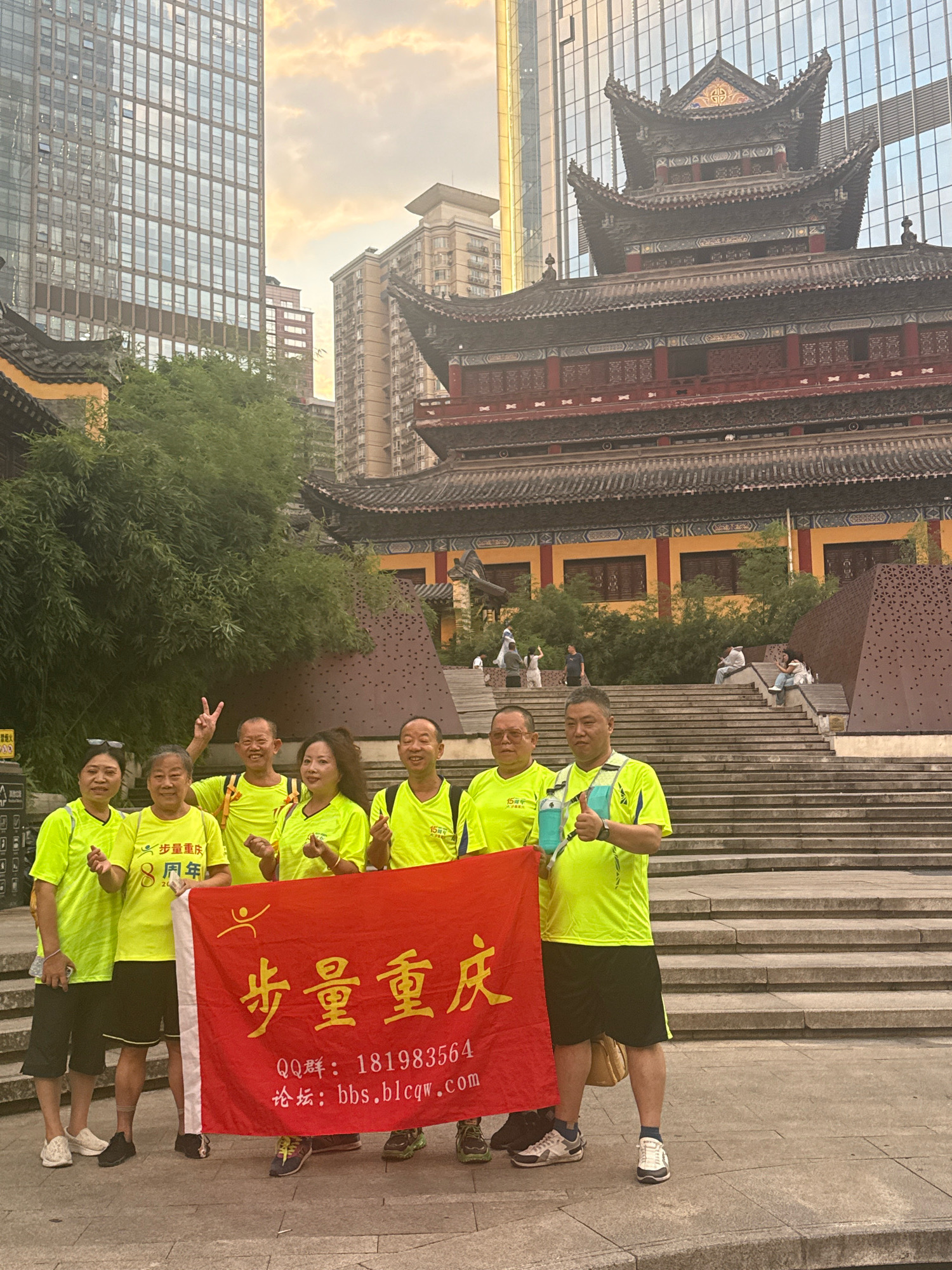 Seven individuals, dressed in bright yellow t-shirts and dark shorts, pose for a photograph in front of a red banner. The banner displays Chinese characters in yellow, with smaller text below. Behind the group, traditional Chinese architecture with a tiered roof dominates the scene, juxtaposed against modern glass skyscrapers in the background, indicating an urban environment. Some individuals are making hand gestures, including a peace sign and a thumbs-up. The group appears to be gathered outdoors on a paved area, with steps leading up to the traditional building visible. The lighting suggests it is either morning or late afternoon. The presence of modern buildings alongside traditional structures suggests the location is likely within a city that has undergone significant development, such as Yuzhong District, China, known for its blend of old and new architecture.