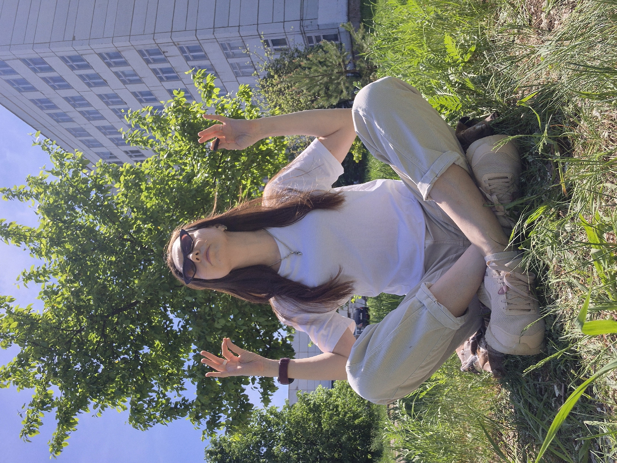 A young woman is sitting cross-legged in a grassy area, possibly a park. She is wearing a white t-shirt, beige pants, and beige sneakers. She also has sunglasses on and a bracelet on her left wrist. Her hair is long and brown. She is holding her hands up, palms open. Behind her are trees and a tall building with many windows. The sky is blue. It is daytime and appears to be sunny. The location is likely urban, suggested by the presence of the building.