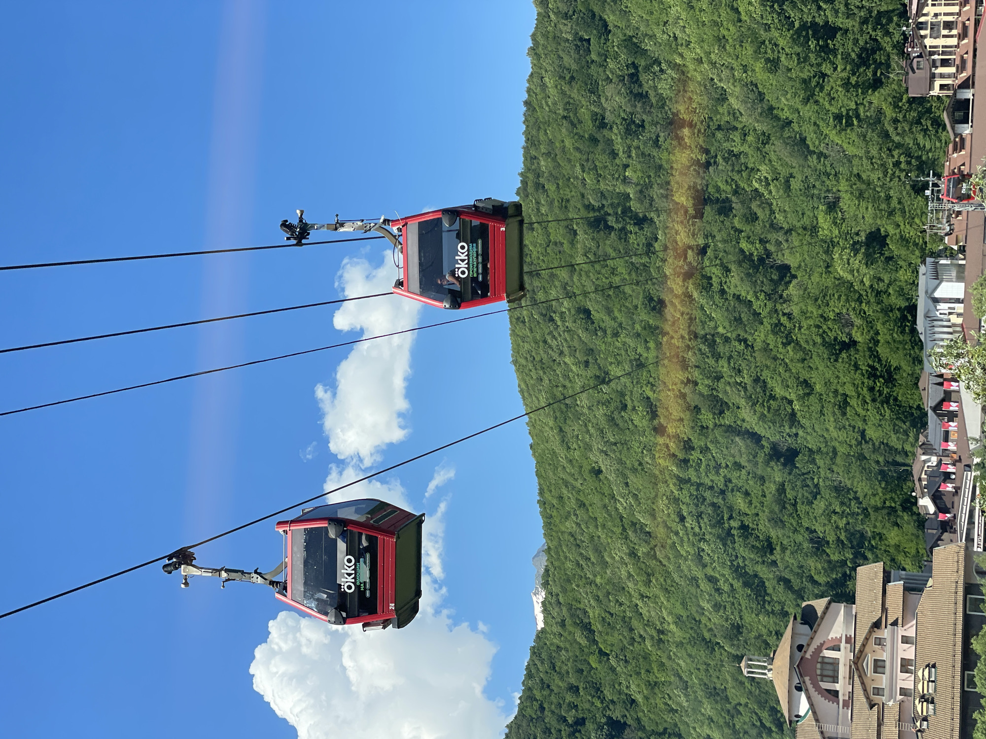 This is a vertical, low-angle shot of two red cable cars suspended on wires against a bright blue sky. The cable cars are positioned one above the other, with the lower one closer to the viewer. Each cable car has large windows and displays the "ökko" logo and Cyrillic text. The scene is set against a backdrop of a lush, densely forested mountain that slopes down towards buildings at its base. A faint rainbow arc is visible on the mountainside, and cumulus clouds dot the sky. The sun appears to be high, suggesting it is daytime, and the weather seems clear and pleasant. The setting suggests a mountainous resort area. The cable cars are likely transporting people up or down the mountain, implying a recreational or transit activity.