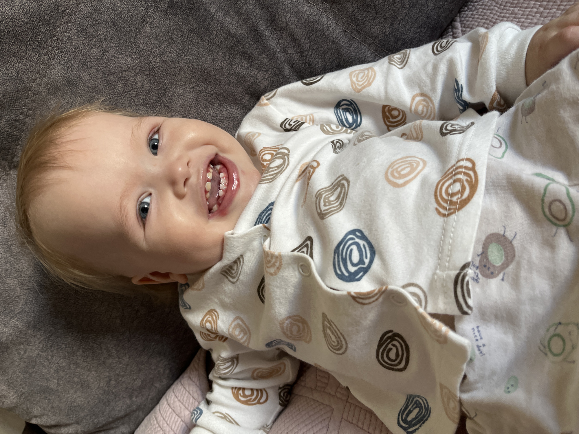 The image depicts an infant situated in an indoor, residential environment within the city of Chaikovsky, Russia. The infant, possessing light skin, light blonde hair, and blue eyes, is observed lying supine on a dark gray, textured upholstered surface. A light pink or purple ribbed fabric is partially visible to the infant's left. The infant is dressed in a two-piece white sleepwear or loungewear set. The long-sleeved top features abstract, spiral-like patterns in shades of brown, dark blue, and tan. The visible portion of the lower garment displays lighter, abstract shapes, and upside-down text that reads, "have a nice day!". The infant is looking directly at the camera with a wide, open-mouthed smile, revealing its upper and lower front teeth. Saliva is present on the lips and gums, and the gums above the upper incisors exhibit a slight reddening.