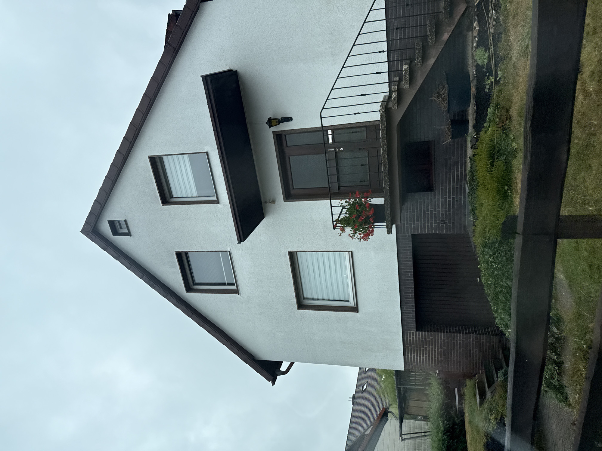 This image shows a residential house in Bad Sachsa, Germany. The house has a white stucco exterior with a dark brown roof and trim. There are several windows on the front of the house, with roller blinds. A set of stairs with a black metal railing leads up to the front door, which is dark brown. Above the door is a small dark brown awning. A light fixture is mounted to the wall next to the door. In front of the door is a potted plant with red flowers. To the left of the entrance, there is a garage with a dark brown door. The property is surrounded by greenery, including bushes and grass. The sky is overcast.