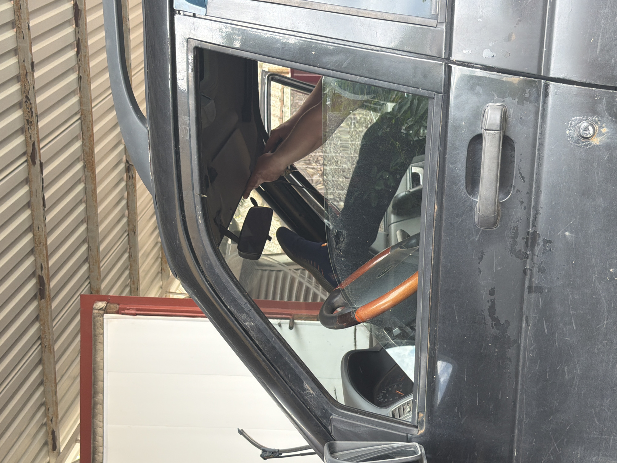 The image shows the partially open door of a dark-colored vehicle, possibly a truck or SUV. A person's hand is visible inside the door frame, appearing to be working on the interior. The steering wheel, dashboard, and part of the driver's leg are also visible inside the vehicle. The vehicle is parked under a corrugated metal roof, likely a carport or garage. Reflected in the glass is a view of greenery. The location and city cannot be determined from the provided image.