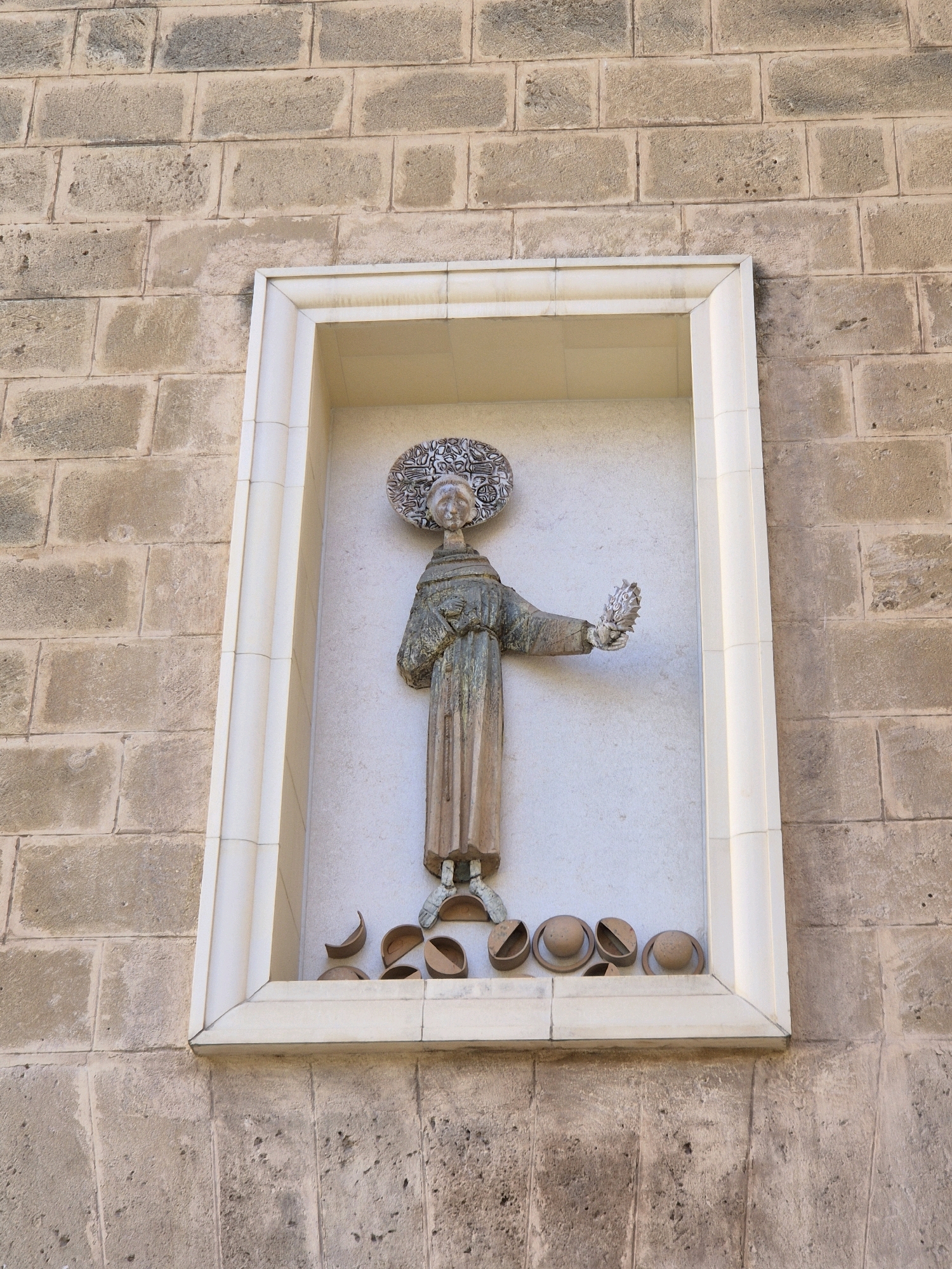 A framed relief sculpture of a saint is mounted on a weathered brick wall. The sculpture depicts a figure in a long robe with a halo around its head, holding a bouquet of flowers in its right hand. The saint's face is serene, and its posture suggests a blessing or offering. At the base of the sculpture, several small, rounded objects resembling bowls or hats are arranged. The wall is constructed of large, rectangular stone blocks, some of which are discolored and worn. The framed sculpture appears to be set into a recessed niche within the wall. The scene suggests a religious or historical site within the city of Taranto, Italy, possibly a church or an ancient building.