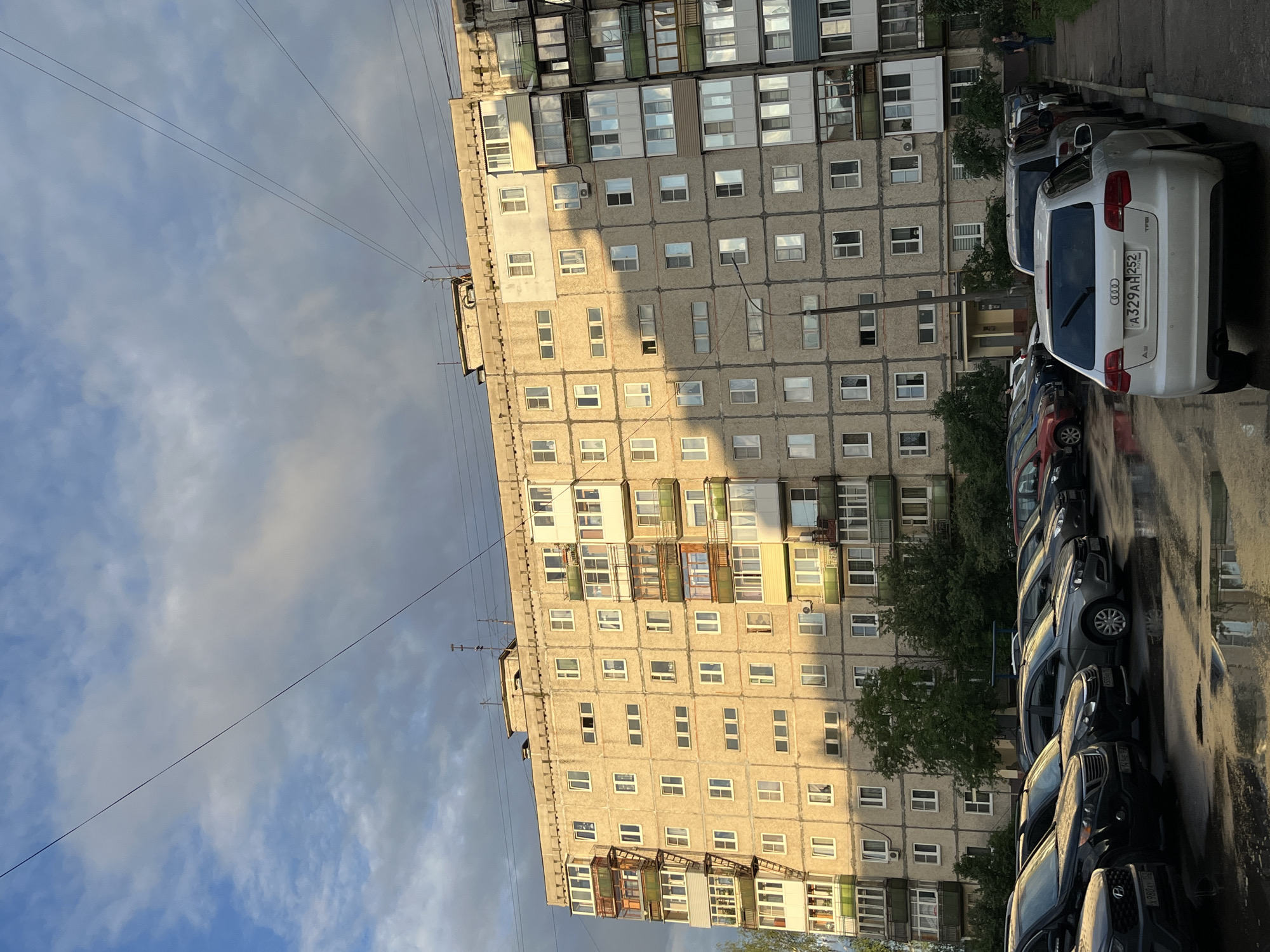 An eye-level, full exterior shot captures a multi-story apartment building with a grid of windows. To the left of the building is a fire escape. In front of the building is a wet parking lot with several cars parked. The sky above is partly cloudy. The image is taken in an urban setting.