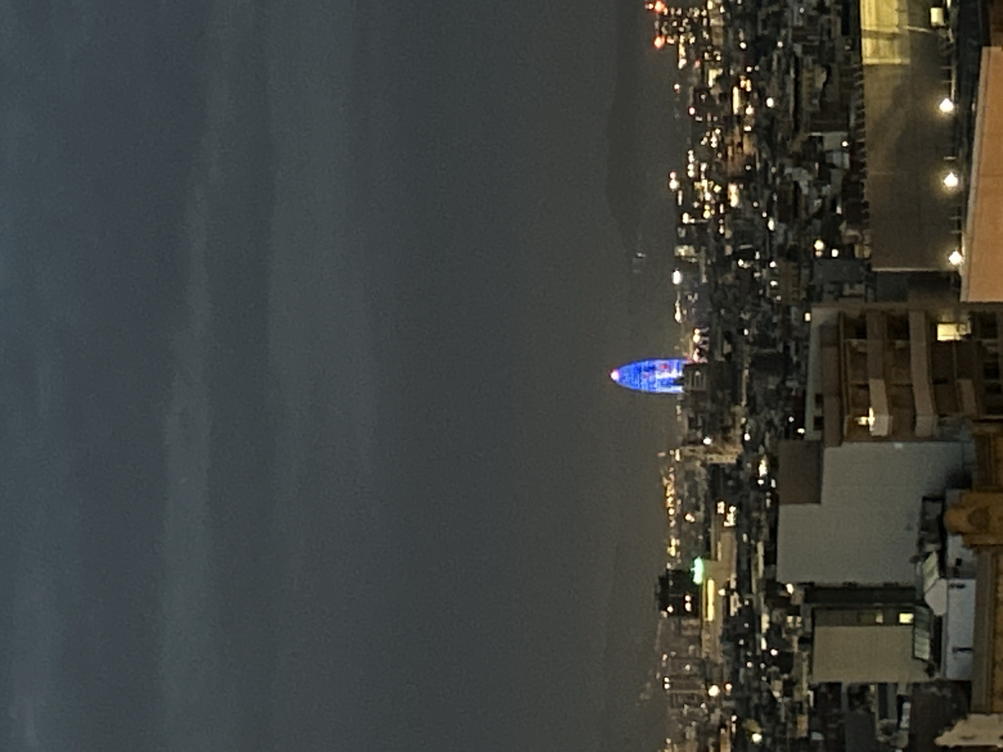 This nocturnal image presents a panoramic view of the Barcelona cityscape from an elevated perspective. The central and most prominent subject is the distinctive, bullet-shaped Torre Glòries (formerly Agbar Tower), illuminated with vibrant blue and purple lights. This iconic skyscraper stands out against the dark night sky in the mid-ground.
To the right of the illuminated tower, the city unfolds into a sprawling expanse of urban lights. Countless golden and white pinpricks of light, emanating from buildings, streets, and homes, create a dense tapestry that stretches across the frame, indicating a lively urban environment. The dark forms of various buildings and structures are visible in the foreground and mid-ground on the right, some with lit windows, hinting at the architectural density of the city.
The sky is dark, confirming the nighttime setting. A faint, darker silhouette of a mountain range or elevated landmass is discernible in the distant background, partially obscured by the darkness. There are no visible people or discernible text within the image. The scene conveys a sense of urban grandeur and the quiet energy of a city after dusk, under clear weather conditions.
