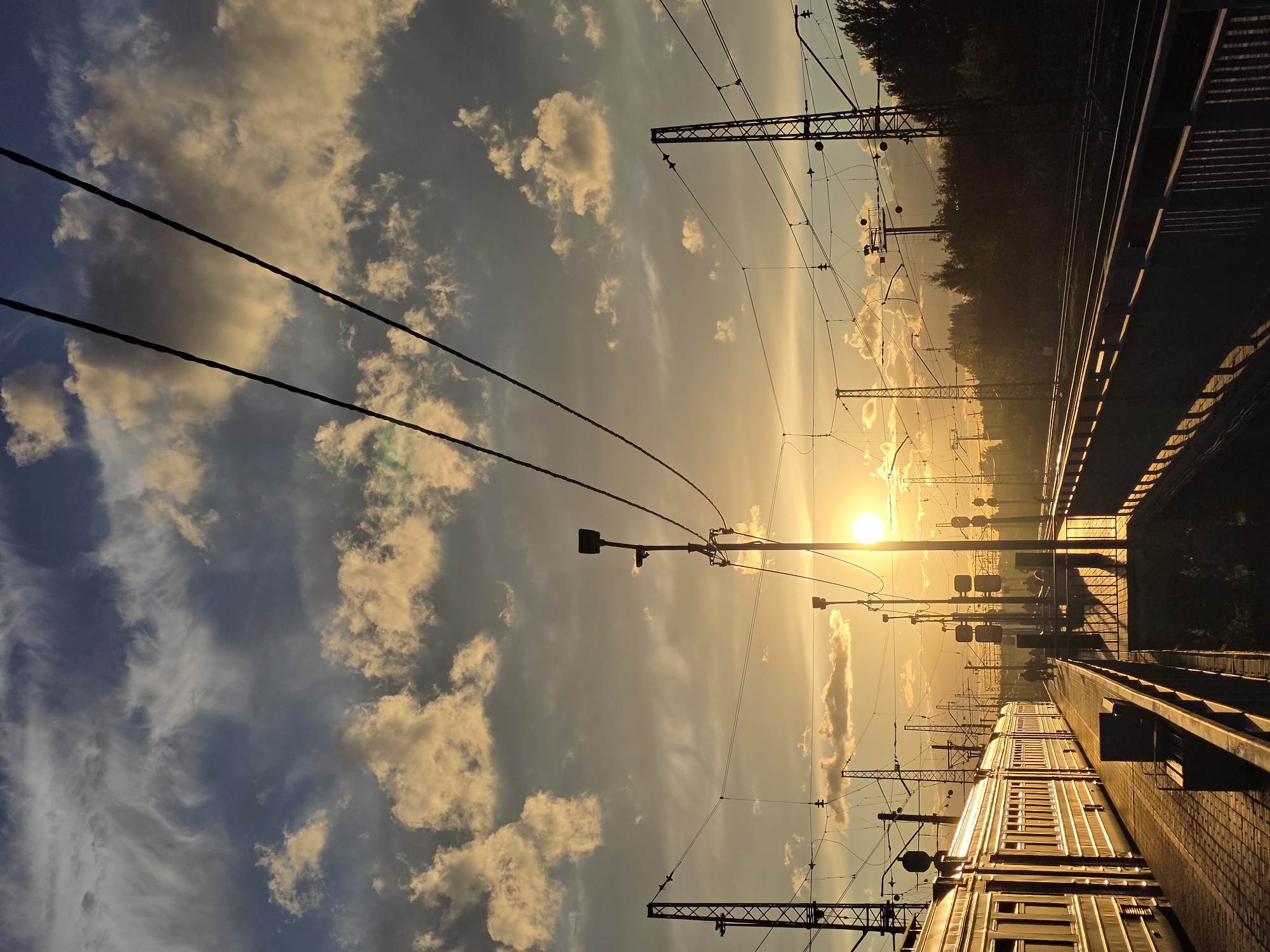 A train station platform is illuminated by the setting sun in Posolok Kombinata Stroymaterialov-2, Russia. A long train with white carriages is stopped on the tracks to the left of the frame, its side reflecting the warm golden light. People are walking along the platform. Overhead, electrical wires crisscross the sky, supported by tall metal poles. The sky is filled with dramatic clouds, some tinged orange and yellow by the sun. Trees and distant buildings are silhouetted against the horizon. The scene conveys a sense of quiet transition as day turns to evening at this suburban train station.