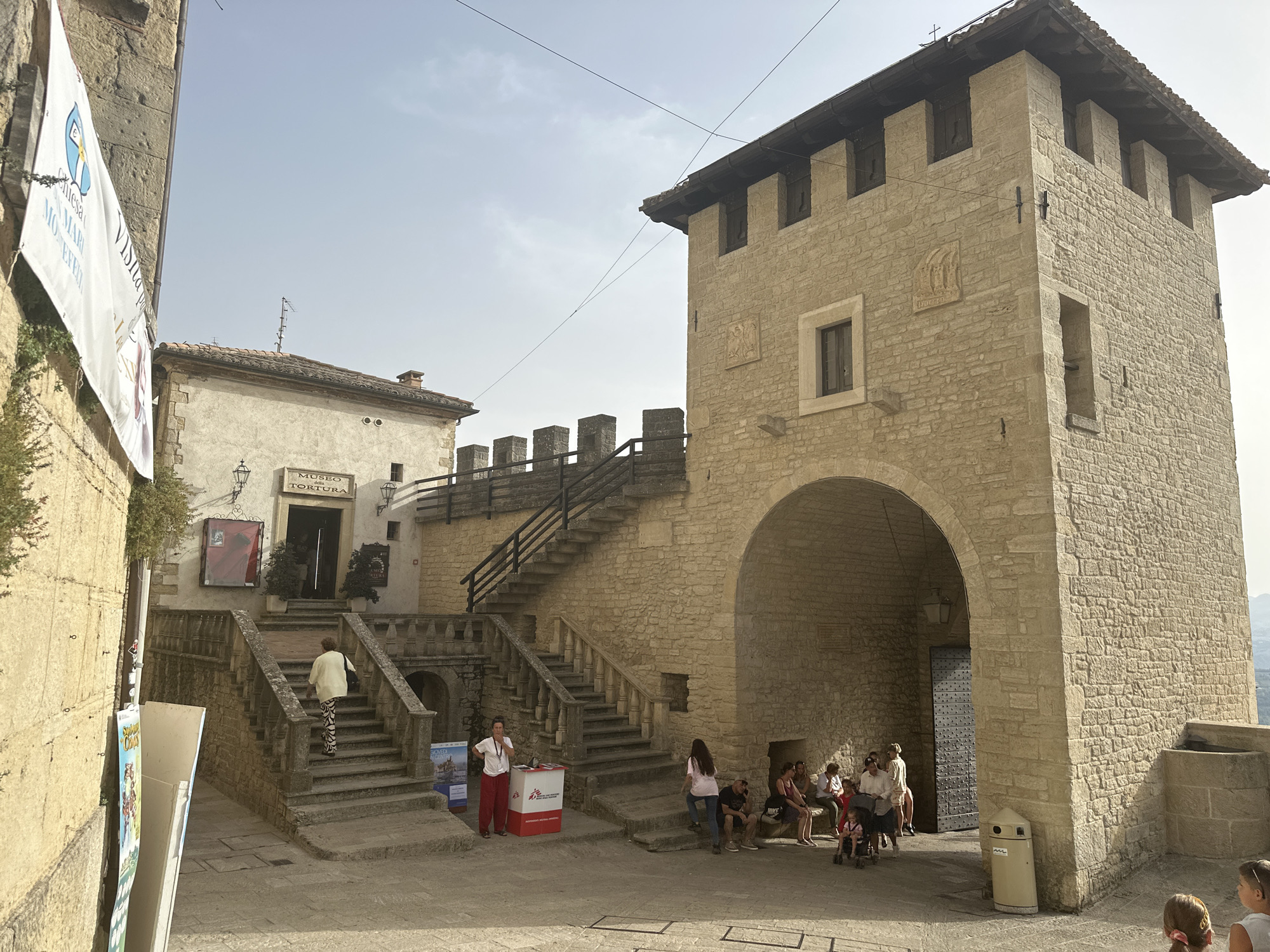 The image shows a view of the City of San Marino, San Marino, featuring the Museum of Torture and a large stone tower with an arched entrance. In the foreground, several tourists are gathered, with some seated on a stone ledge and others standing or walking. A woman in red pants and a white shirt stands next to a red and white kiosk. A woman in a yellow jacket is walking up a stone staircase leading to the museum entrance. A banner with text and a logo is attached to a stone wall on the left side of the image. The sky is clear and bright.