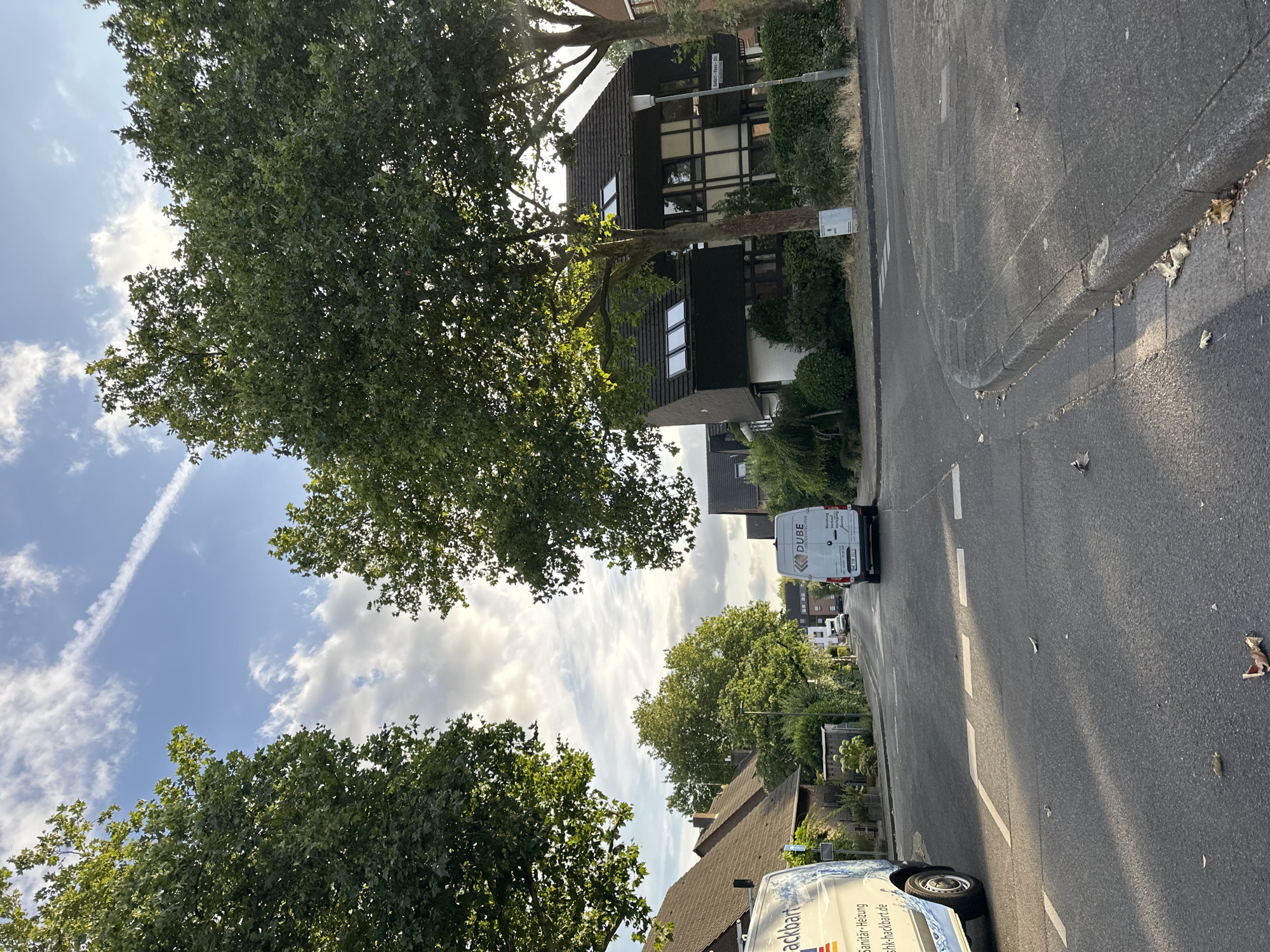 The image depicts a residential street in Krefeld, Germany, with mature trees lining the road. Two white commercial vans are visible. One van on the left has text in German that appears to be "Sanitär-Heizung" and a website address. The van on the right displays "DUBE FUßBODENTECHNIK" and other German text. The street has asphalt pavement with white lane markings and a sidewalk. Buildings with pitched roofs are visible on the right side of the street. The sky is blue with scattered clouds and a contrail. The lighting suggests it is daytime, possibly late afternoon or early evening, with sunlight filtering through the trees. There are no people clearly visible in the image.