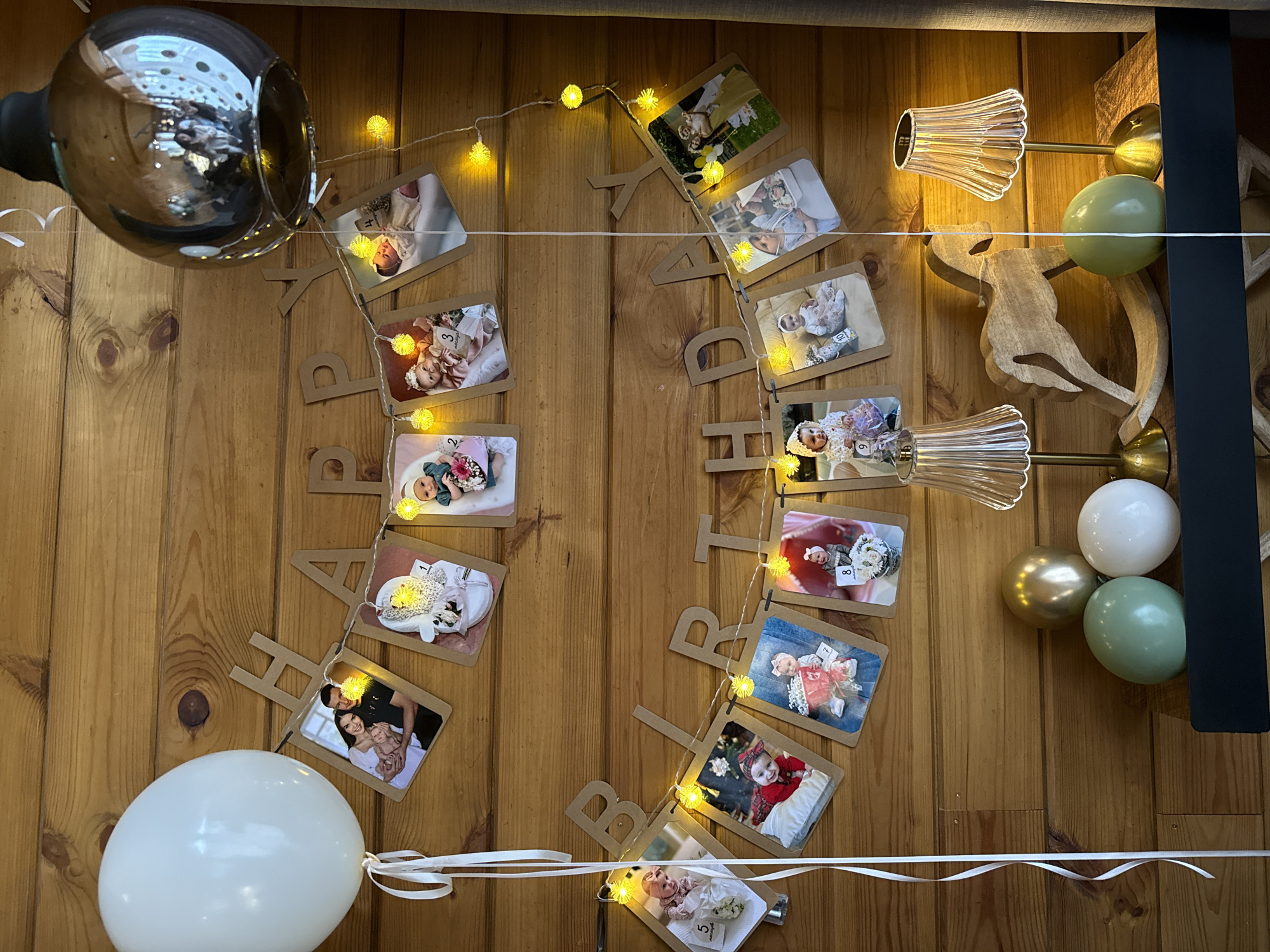 This is a medium shot of a "Happy Birthday" banner with photos and fairy lights strung on a wooden wall. The banner is composed of individual letter cutouts, and interspersed between them are small framed photographs of a baby. Each photo has a number overlaid on it, indicating the baby's age in months. There are also several balloons visible: a large white balloon on the left, and a cluster of three smaller balloons (gold, light green, and white) on the right. Two glass sconces with ribbed shades are mounted on the wall, providing ambient lighting. A large, reflective spherical object is partially visible in the top left corner. The scene suggests a birthday celebration, likely for a baby given the photographs. The overall mood is festive and celebratory.