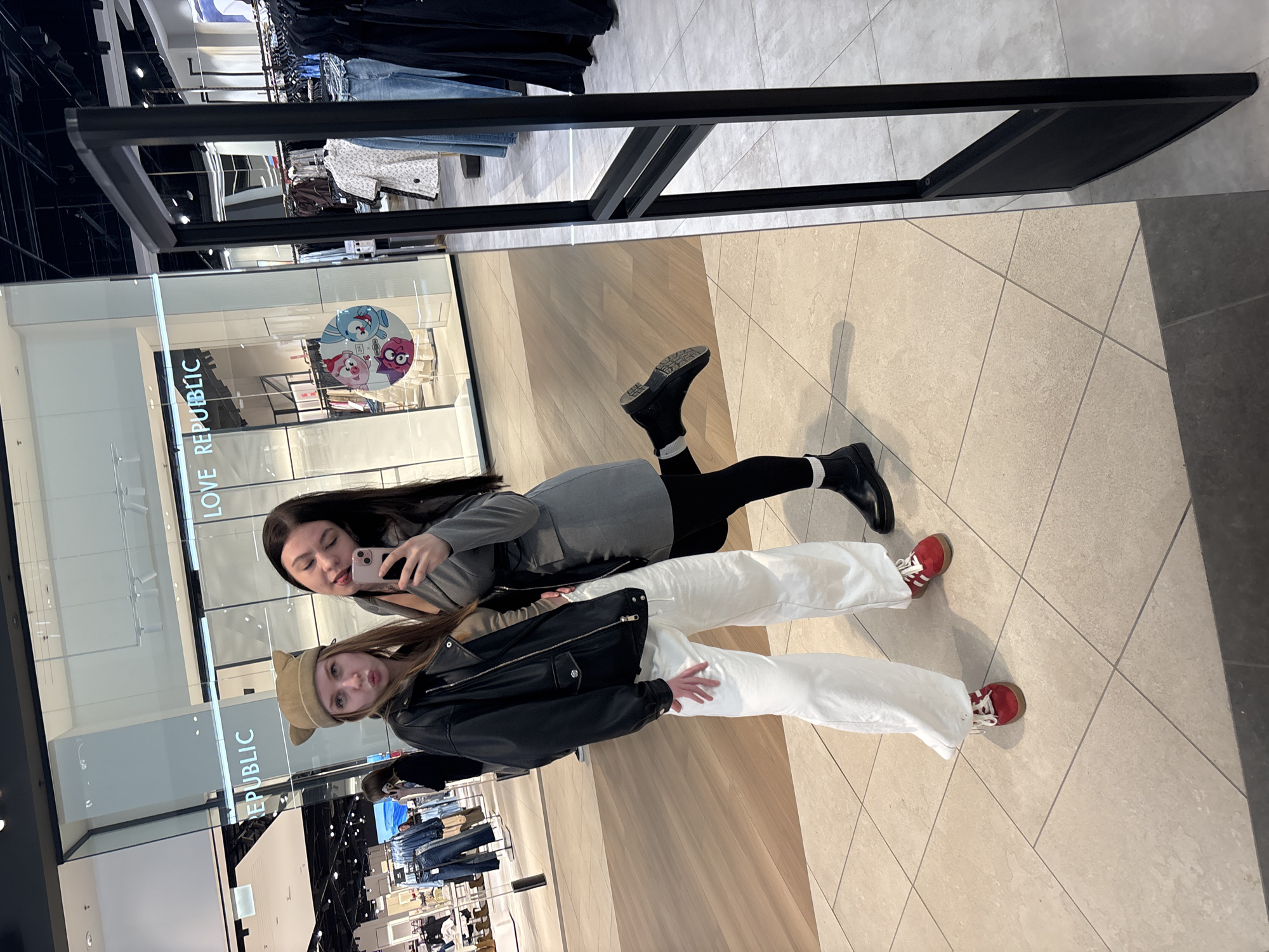 Two young women are taking a selfie in a shopping mall. One woman wears a grey top, black leggings, and black boots with white socks. The other woman wears a beige hat, a black jacket, and white pants with red shoes. They are posing in front of a store named "LOVE REPUBLIC". The mall has a light-colored tile floor. Clothing racks are visible in the background. Based on the store name and general context, this is likely in the city of Perm, Russia.