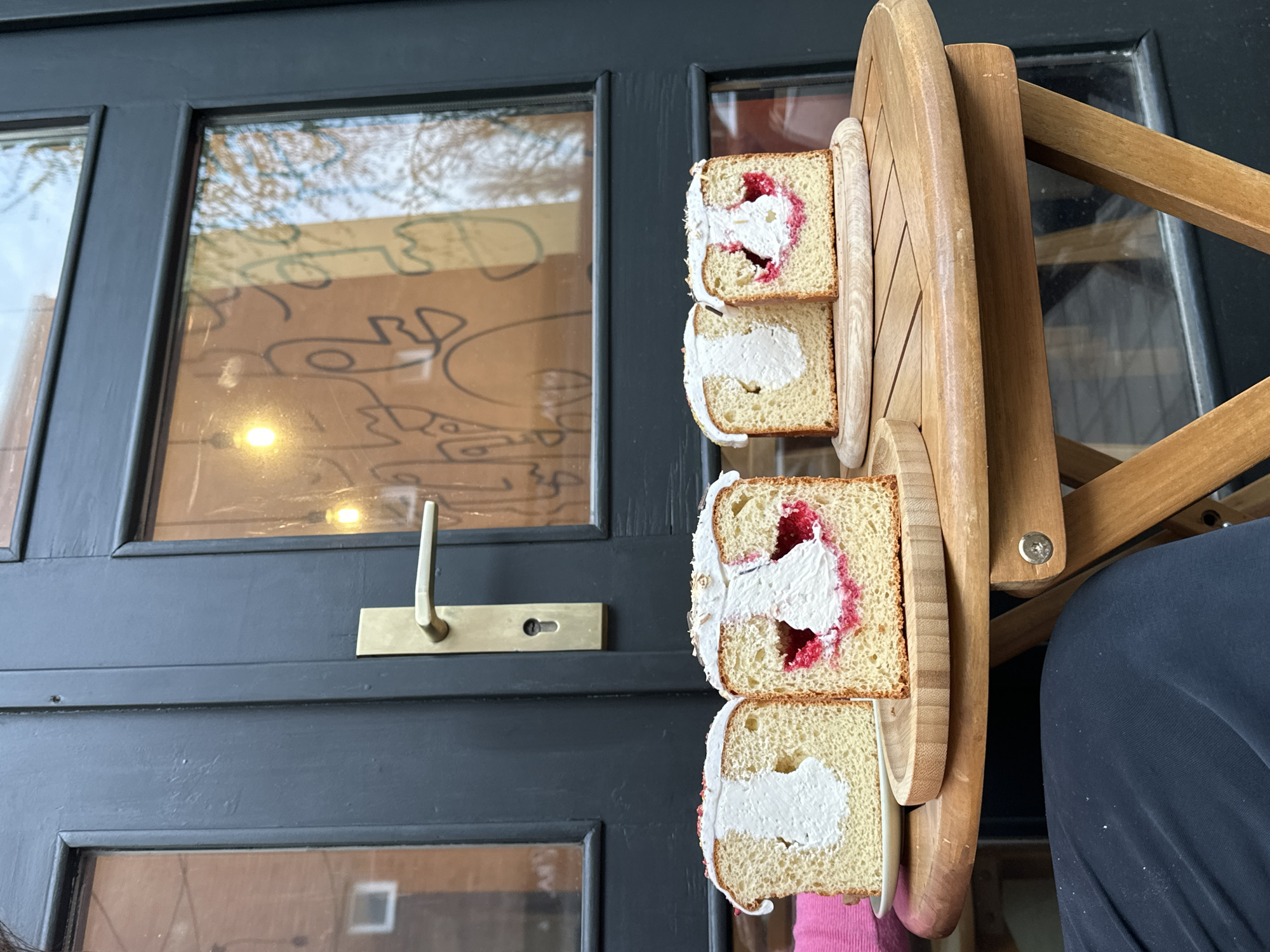 The image shows four square slices of cake, each containing a white cream filling and red jam or fruit filling, presented on a wooden multi-tiered serving stand. The cake slices are stacked vertically, with two slices per tier. The serving stand is placed in front of a black door with glass panels. The door has a brass handle and keyhole. The reflections in the glass panels suggest an outdoor environment, including trees and a building with a painted design. The location could be a cafe or restaurant patio in Novi Sad, Serbia. A portion of a wooden chair and a person's clothing (dark blue fabric) are visible on the right side of the image.