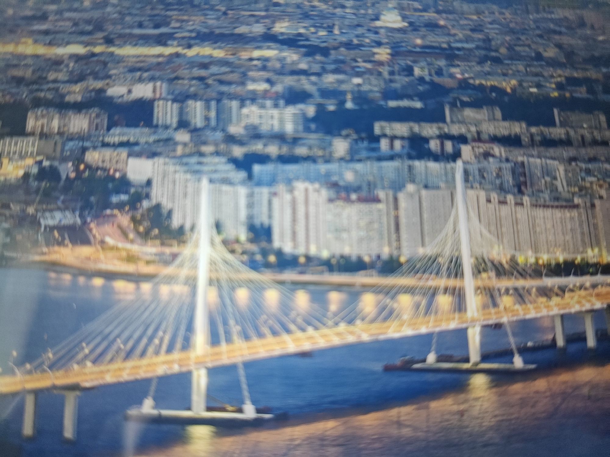 The image depicts a panoramic view of a city skyline at dusk, with a prominent cable-stayed bridge spanning a body of water in the foreground. The bridge is illuminated by warm lights, casting reflections on the dark blue water. In the background, a dense urban landscape with numerous multi-story buildings stretches towards the horizon. The buildings appear to be residential and commercial structures. The sky is a muted blend of blues and oranges, suggesting the transition from day to night. The overall impression is one of urban development and infrastructure.