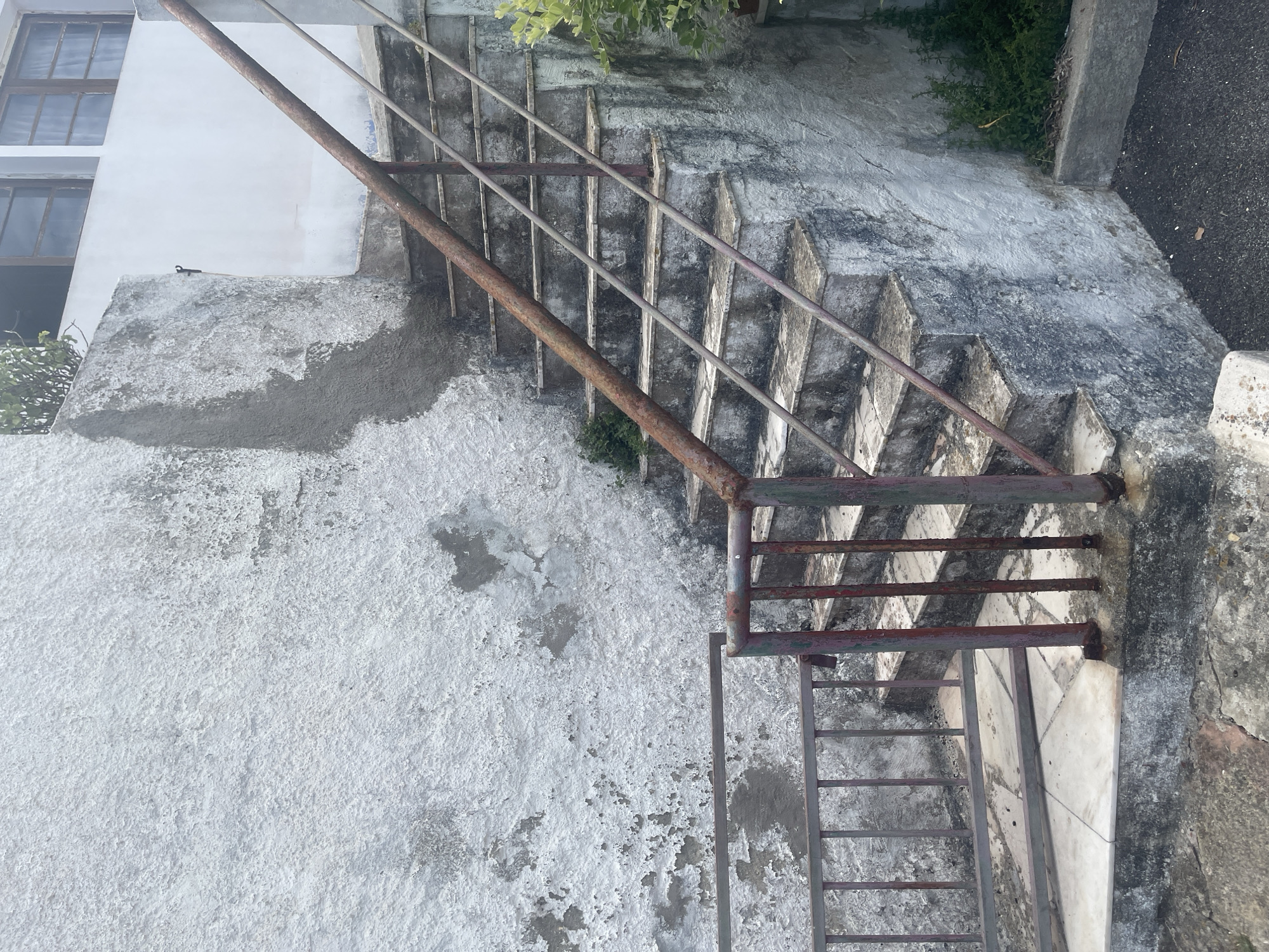The image depicts an outdoor staircase with a rusty railing, set against a weathered, white-painted concrete wall. The wall shows signs of chipping and discoloration, with patches of exposed concrete and darker stains. The stairs themselves are made of concrete with white tiled risers and are also weathered and stained. Greenery, consisting of small bushes, is visible growing near the base of the stairs and along the side of the wall. The foreground shows a concrete pathway and a section of a metal railing. The scene appears to be in an urban or suburban environment, likely in Colares, Portugal, given the architectural style and the presence of Mediterranean-style vegetation. The lighting suggests an overcast day. There are no people visible in the image.