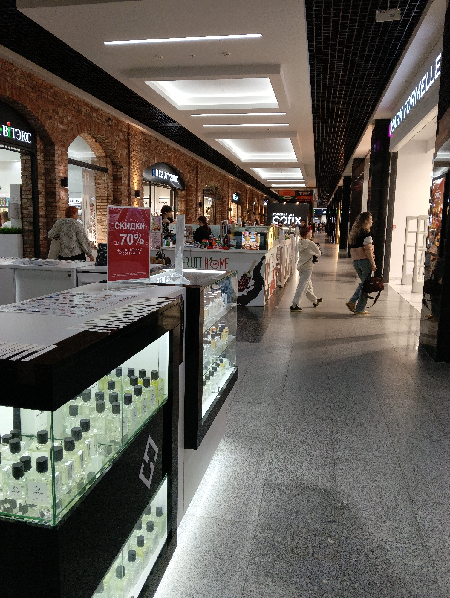 The image depicts a well-lit indoor shopping mall corridor, likely in the city of Hrodna, Belarus, given the Russian text on signs. The corridor features a prominent display of perfumes and cosmetics in glass cases on the left side of the frame. A red sign with Cyrillic text, indicating "discounts up to 70%", is prominently displayed on a stand.
Several individuals are present:
- In the foreground, a woman with short, reddish-brown hair and a patterned shirt is facing away from the camera, interacting with a display.
- Further down the corridor, two women are walking away from the camera. The one on the left wears a light-colored tracksuit, and the one on the right wears a dark top and blue jeans, carrying a brown bag.
- In the background, other shoppers and staff are visible near shop entrances.
The environment includes:
- Archways and brickwork on the left side of the corridor, suggesting a stylized architectural design.
- Suspended ceilings with integrated lighting.
- Shop fronts with signage, including "BEAUTYZONE" and "MARK FORMELLE."
- A polished gray tiled floor.
- A glass display counter filled with numerous small bottles, likely perfumes, with black caps.
The overall scene suggests a commercial setting with active shopping activity.