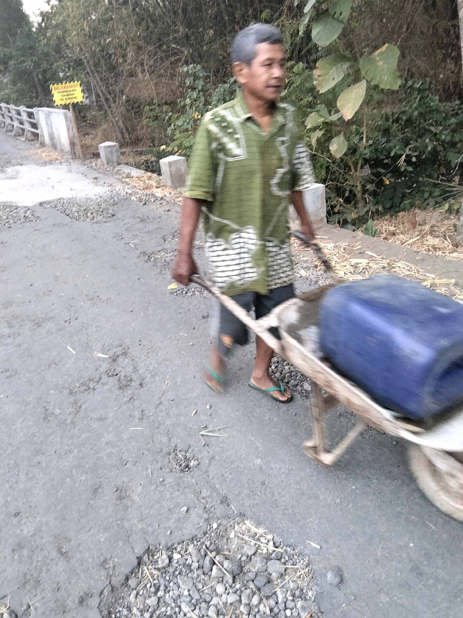 A man is pushing a wheelbarrow containing a large blue plastic container along a paved road in Jenangan, Indonesia. The man is wearing a green and white checkered shirt, dark shorts, and flip-flops. The road is cracked and uneven, with gravel scattered on the surface. In the background, there is vegetation and a yellow sign with Indonesian text. The scene appears to be outdoors during daylight.