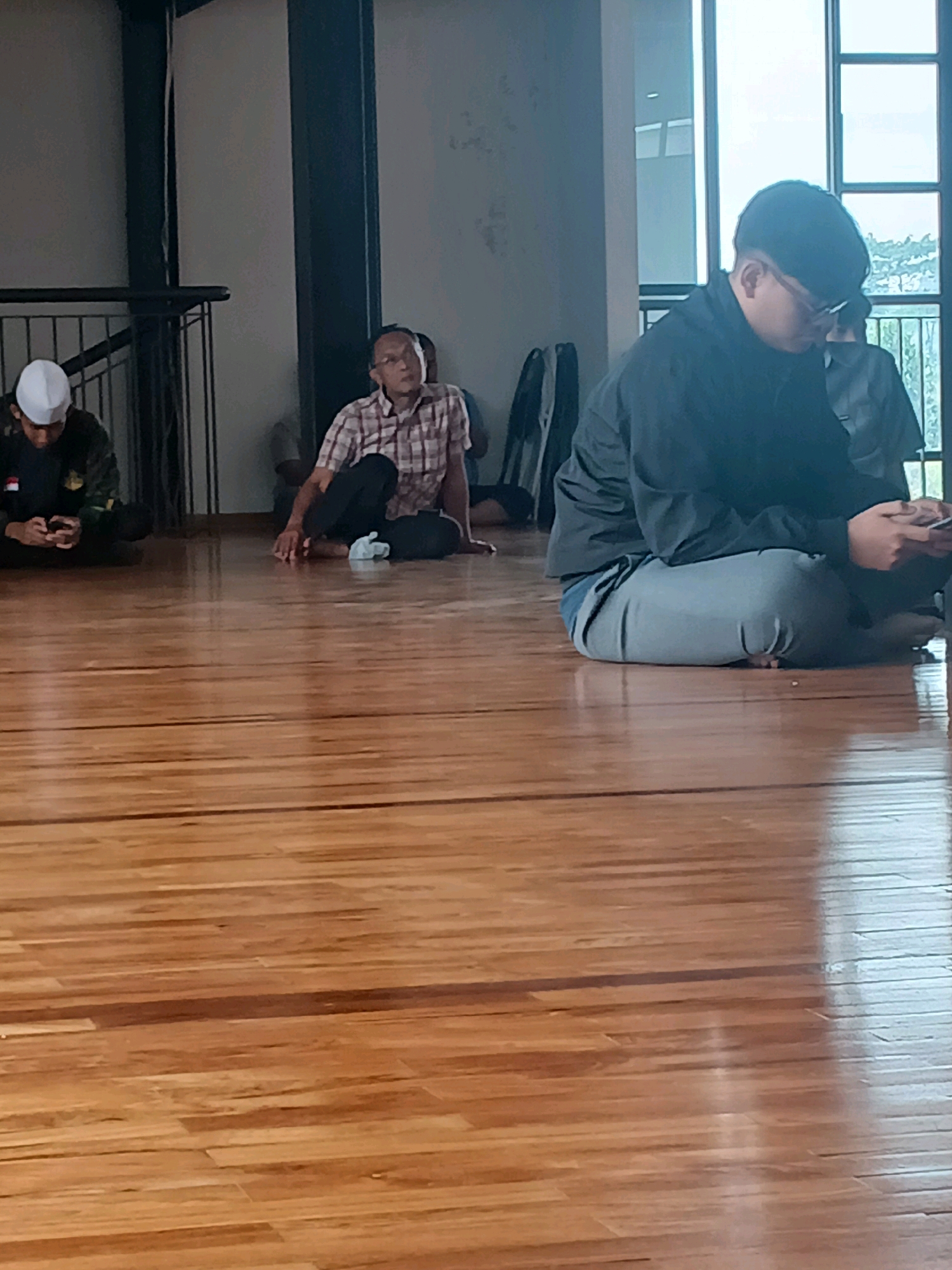 In a brightly lit indoor space with a polished wooden floor, four individuals are seated on the floor. The individual on the far left wears a white skullcap and a dark uniform with patches, and appears to be looking at a mobile phone. To their right, a man in a plaid shirt and dark pants sits with his legs crossed, looking upwards. Behind him, another person is partially visible. On the right side of the frame, a young man wearing glasses and a black jacket is seated, looking down at a mobile phone. The background consists of a light-colored wall and large windows that offer a view of greenery and a partially visible structure. The environment suggests a communal gathering or a place of worship. The image was captured in Arjasari, Indonesia.