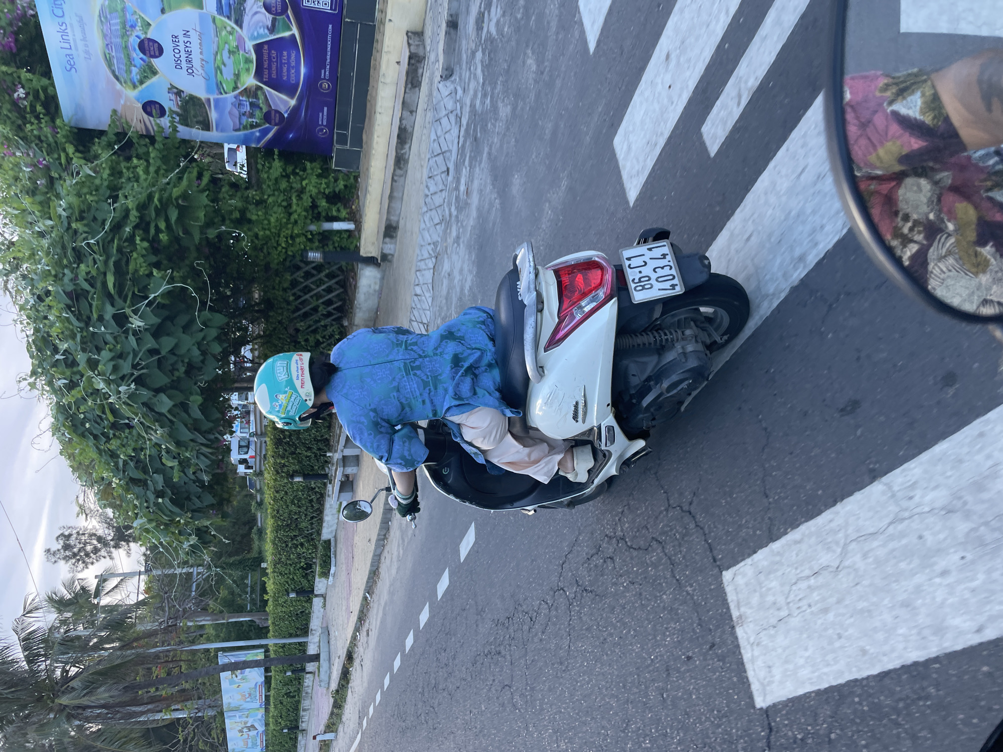 A person on a white scooter rides across a pedestrian crossing on a street in Phu Thuy Ward, Vietnam. The individual is wearing a blue patterned shirt, light-colored pants, and a teal helmet. The scooter has a license plate that reads "86-C1 403.41". In the background, a large billboard advertising "Sea Links City" is visible. Palm trees and lush green foliage line the street, with a cloudy sky overhead. The road surface is asphalt with visible cracks.