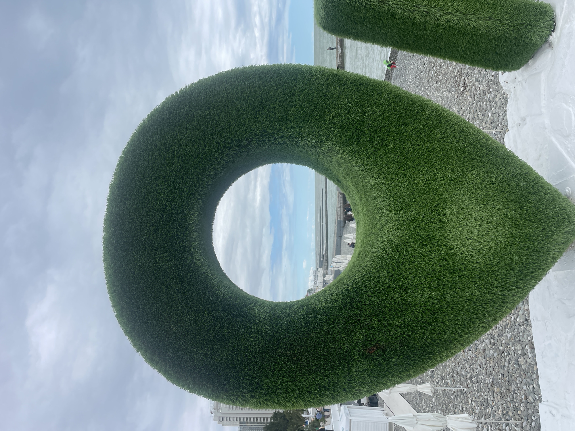The image captures a large, three-dimensional green marker symbol constructed from artificial turf, positioned near a beach in Sochi, Russia. The marker's shape resembles a map pin, with a circular opening in the upper part framing a view of the sea and a cloudy sky. In the distance, figures are visible on a pier extending into the sea.
The foreground includes a pebble beach and white structures, possibly part of a beachfront establishment. A tall building is partially visible in the background, indicating an urban setting. The sky is overcast, contributing to the image's soft lighting. The overall scene suggests a tourist location or public art installation.