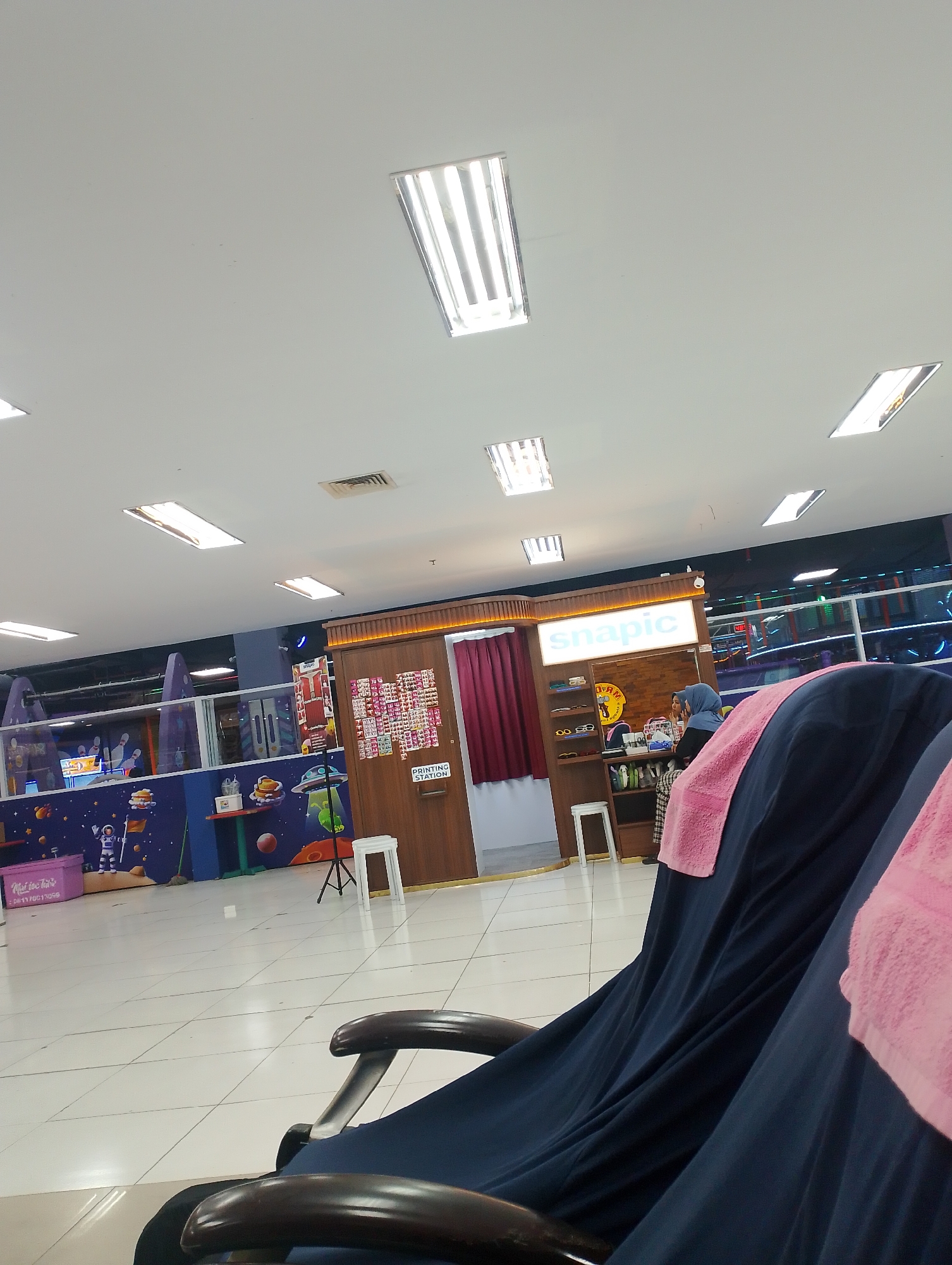 The image depicts the interior of a shopping mall in Batam, Indonesia, specifically in the Nagoya Hill Shopping Mall. In the foreground, two chairs draped with dark blue fabric and pink towels are visible. Beyond the chairs, a "Snapic" photo booth and printing station is set up. A woman wearing a blue hijab is standing behind the counter. The background features a colorful children's play area with a space-themed mural, including astronauts and rockets. Fluorescent lights illuminate the ceiling.
