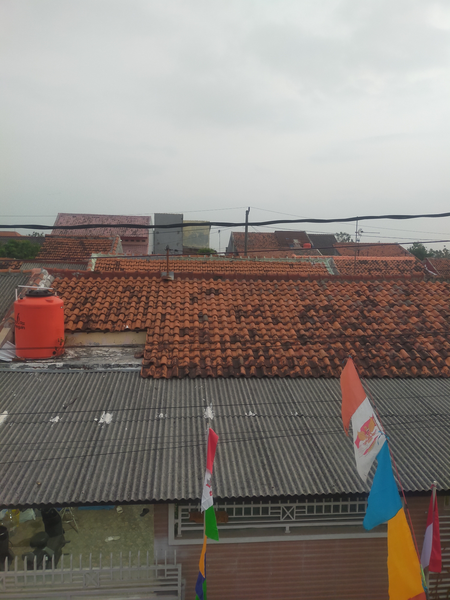 The image shows a view of rooftops and a cloudy sky in Tegalandong, Indonesia. The foreground features a corrugated metal roof with a large, orange water tank on it. Behind this, several other rooftops are visible, primarily covered with traditional terracotta tiles. In the distance, more buildings and some greenery can be seen. Several Indonesian flags, some with the Garuda Pancasila emblem, are flying from poles, suggesting a festive or patriotic occasion. The overall environment appears to be a residential area with a somewhat overcast sky, indicating potentially humid or rainy weather. There are no people visible in the scene.
