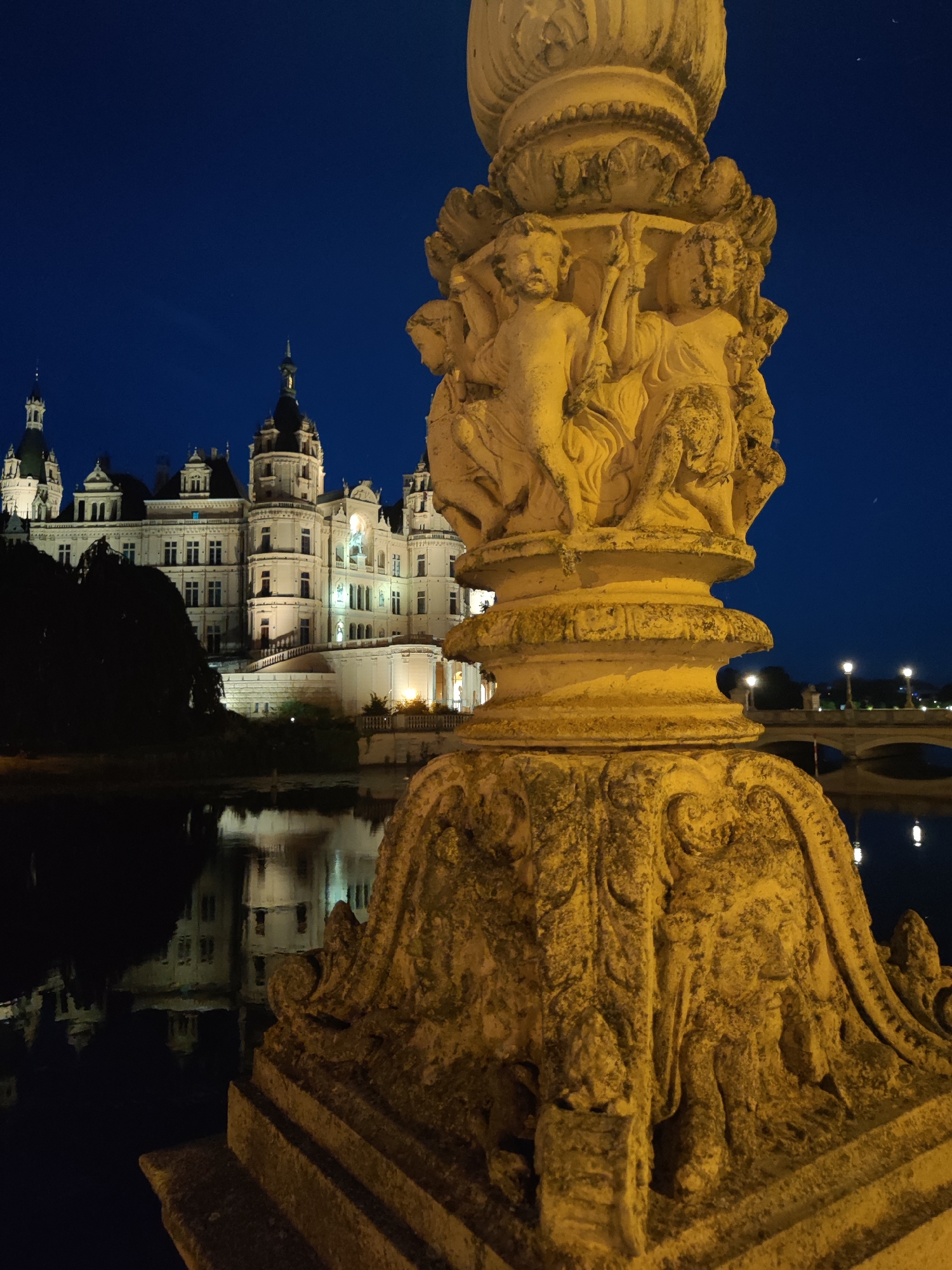 The image shows a view of Schwerin Castle at dusk, with a stone pillar in the foreground. The castle is a large, white building with multiple towers and turrets, illuminated by artificial lights. It is reflected in the dark water in front of it. The stone pillar is ornate, with carved figures and decorative elements. The sky is a deep blue, indicating the late hour. In the background, a bridge with lampposts can be seen spanning the water. The scene is set in Schwerin, Germany.
