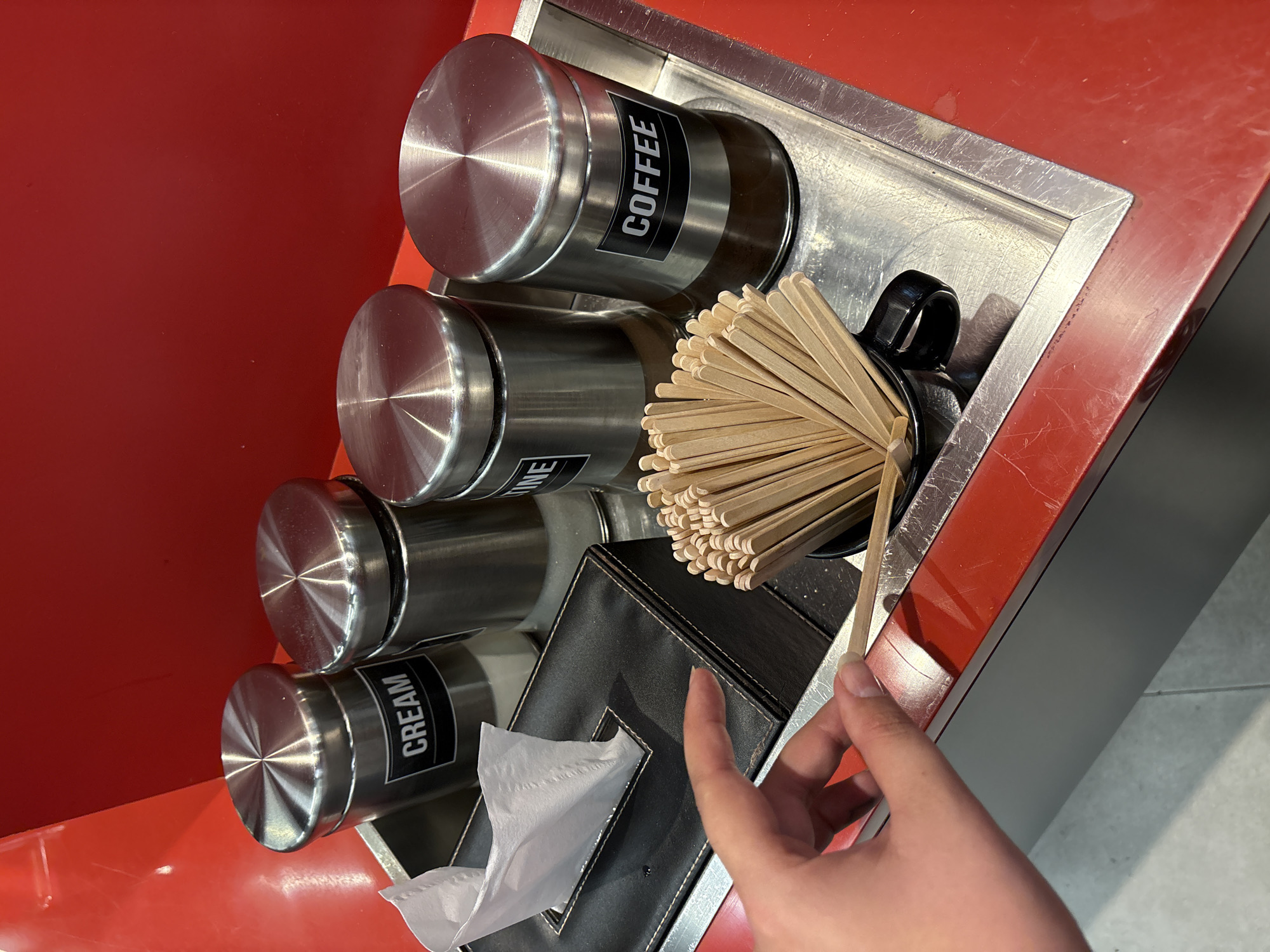 This media file shows a close-up view of a coffee condiment station, likely in a cafe or self-serve area. The main objects are four stainless steel canisters with black labels. The visible labels read "COFFEE" and "CREAM". Another canister's label is partially visible, appearing to say "SUGAR". The fourth canister's label is not visible. These canisters are arranged in a row within a metal tray, which is set against a red surface.
To the right of the canisters, a black mug is filled with a large bundle of wooden stir sticks. A person's hand, with manicured fingernails, is reaching into the mug to take a stir stick. Below the mug and stir sticks, a black leather tissue box holder is visible, with white tissues protruding from it. The overall setting appears to be indoors, possibly a cafe in Nong Pa Khrang, Thailand, given the location context. The lighting suggests it could be daytime. There are no strong emotional cues, but the scene is practical, related to preparing coffee.