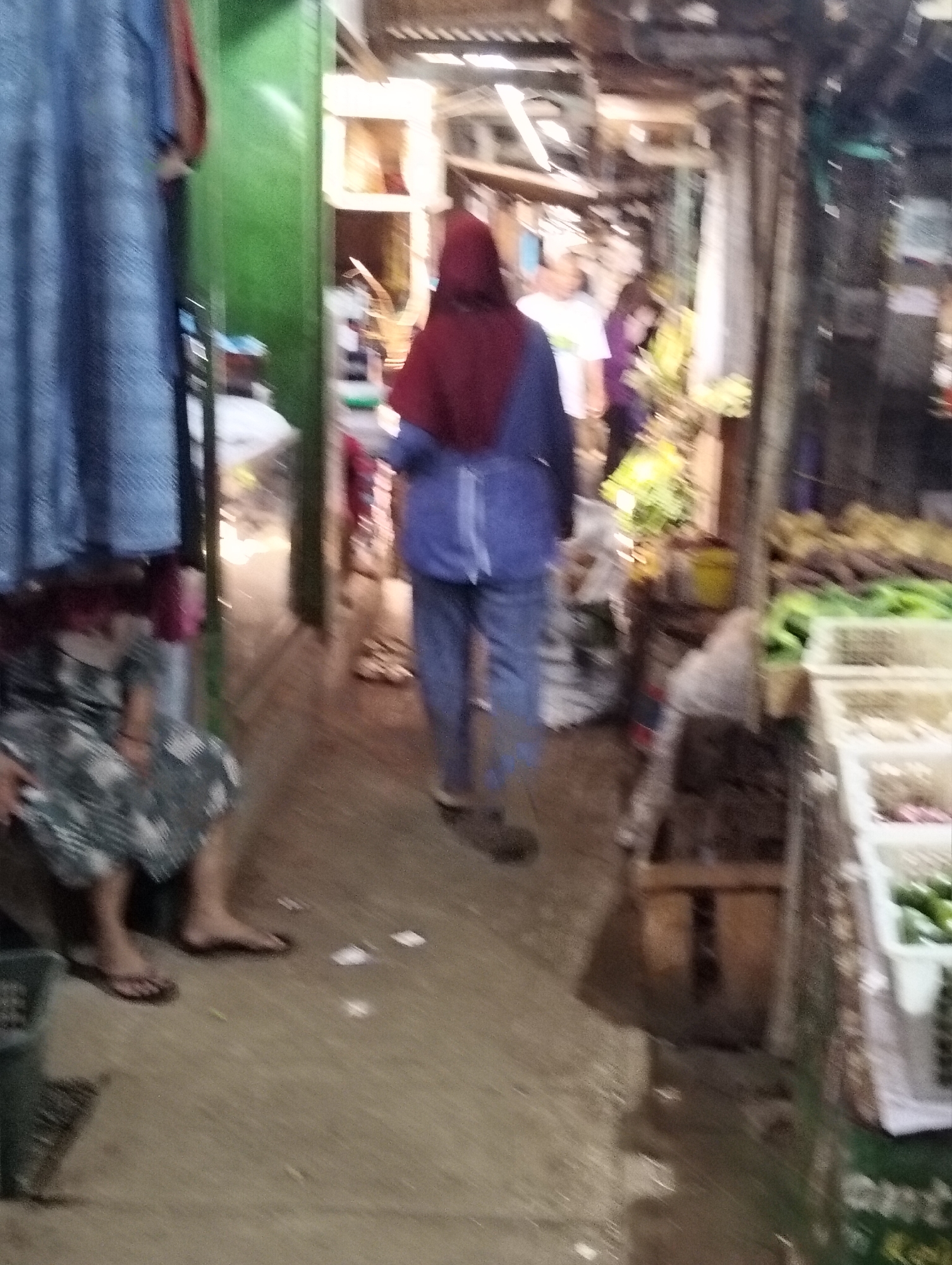 The image depicts a bustling marketplace scene, likely within the city of Subah, Indonesia, characterized by its vibrant and somewhat chaotic atmosphere. In the foreground, to the left, a person is seated, their legs visible, wearing a patterned dress and flip-flops. Their presence suggests a moment of rest or interaction within the market.
Dominating the mid-ground is a person walking away from the camera, their figure partially obscured. They are wearing a maroon headscarf and a blue jacket, with dark pants. This individual appears to be navigating through the market, possibly heading deeper into the stalls or towards an exit.
The environment is a typical market setting, with stalls and products lining the pathway. To the right, baskets filled with green vegetables, possibly cucumbers or similar produce, are visible, indicating the sale of fresh goods. The overall impression is one of a lively marketplace with people engaged in shopping or preparing to sell their wares. The lighting suggests it might be either early morning or late afternoon, with shadows indicating the direction of light sources. The ground appears to be a dirt or concrete pathway, common in such open-air markets. The background is filled with indistinct shapes of stalls, goods, and other people, contributing to the sense of a busy and active commercial hub. The presence of what appears to be tarpaulin or corrugated metal roofing overhead suggests a partially covered market.