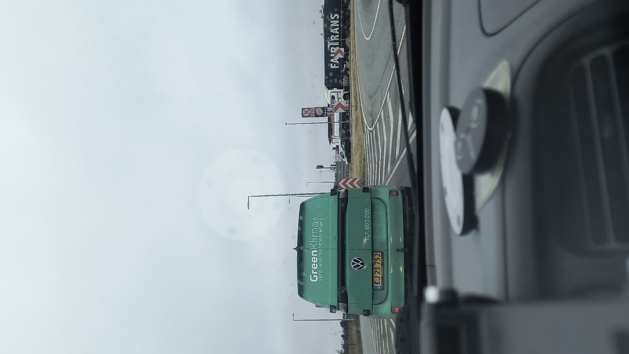 The image was taken from inside a vehicle on a cloudy day in Ølby, Denmark. The vehicle is stopped at an intersection, with a green Volkswagen van in front. The van has a license plate that reads "EJ 25 752" and the words "Green Klima" and "Ventilationsentreprenør" are visible on its rear door, along with a phone number "TLF: 8173 0199". To the right of the van, there is a circular intersection with a large tanker truck that says "FAIRTRANS". In the background, a Burger King and a Circle K gas station are visible. The dashboard of the vehicle in the foreground shows two round objects, possibly car remotes or key fobs.